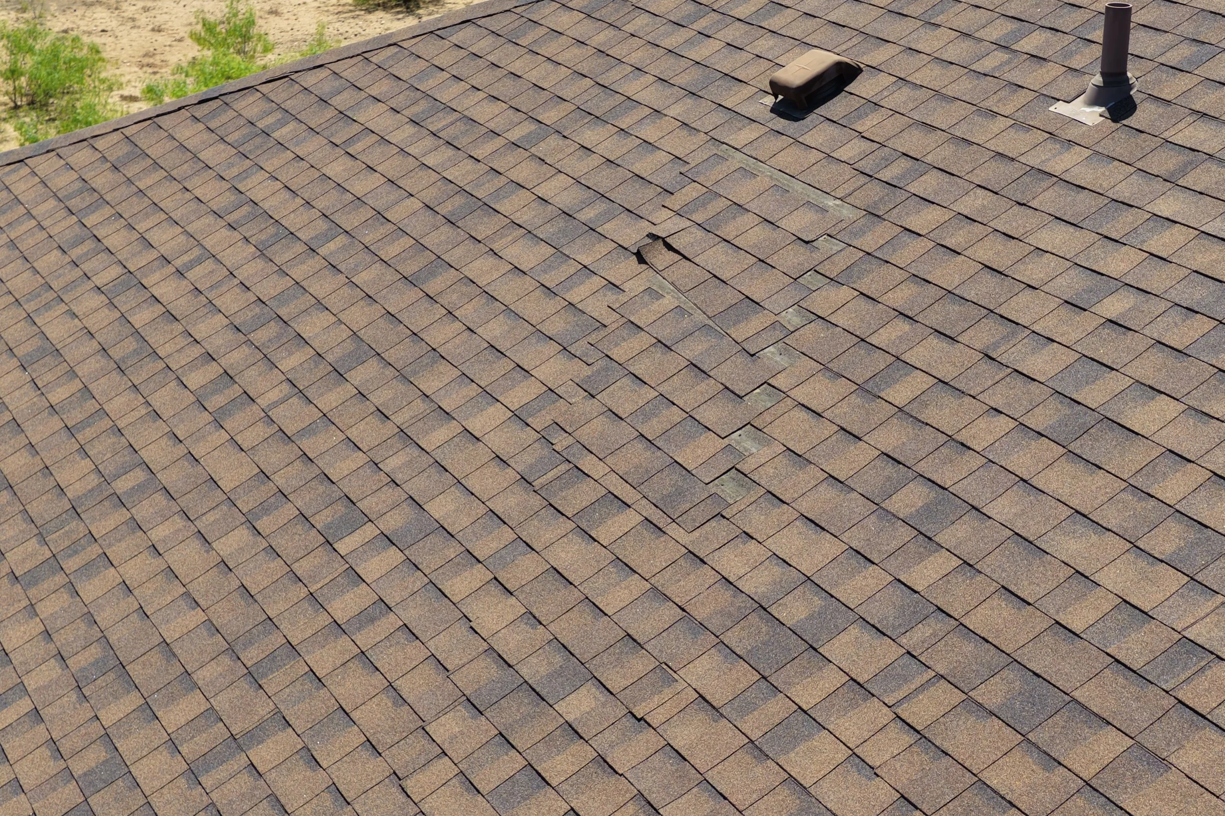 Close-up of a brown shingle roof with two vents, one flat and rectangular, and the other cylindrical. Some shingles are slightly lifted.