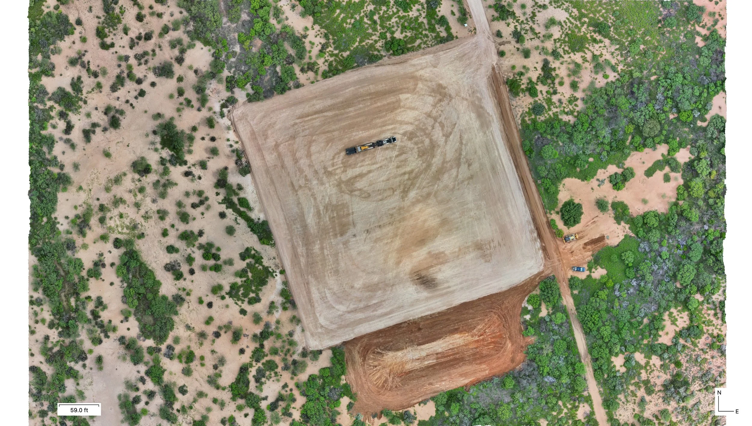 Aerial view of a construction site with cleared land and a dirt road, surrounded by green trees and shrubs.