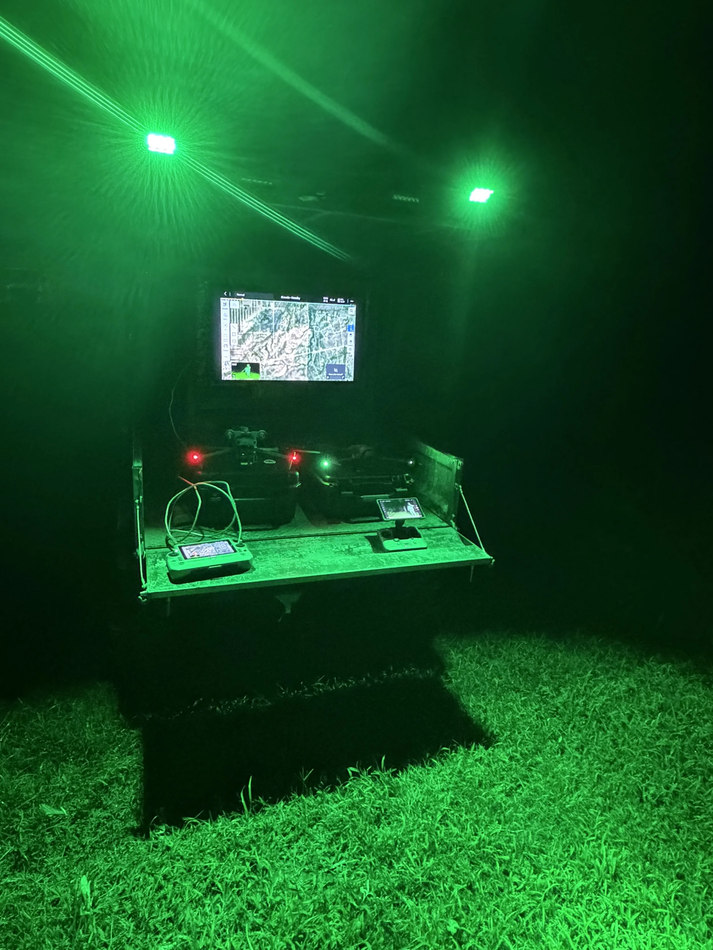 A nighttime outdoor scene with green laser lights casting beams into the dark sky. A table with electronic equipment, including monitors and drones, is set up on the grass, illuminated by the green laser beams.