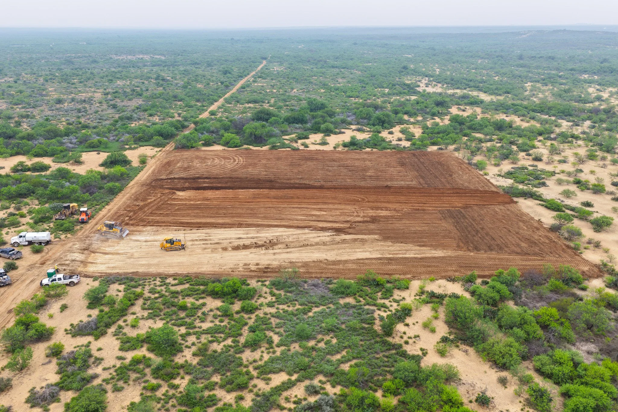 Aerial view of a landscape with a section of cleared land and construction equipment, surrounded by green trees and shrubs.