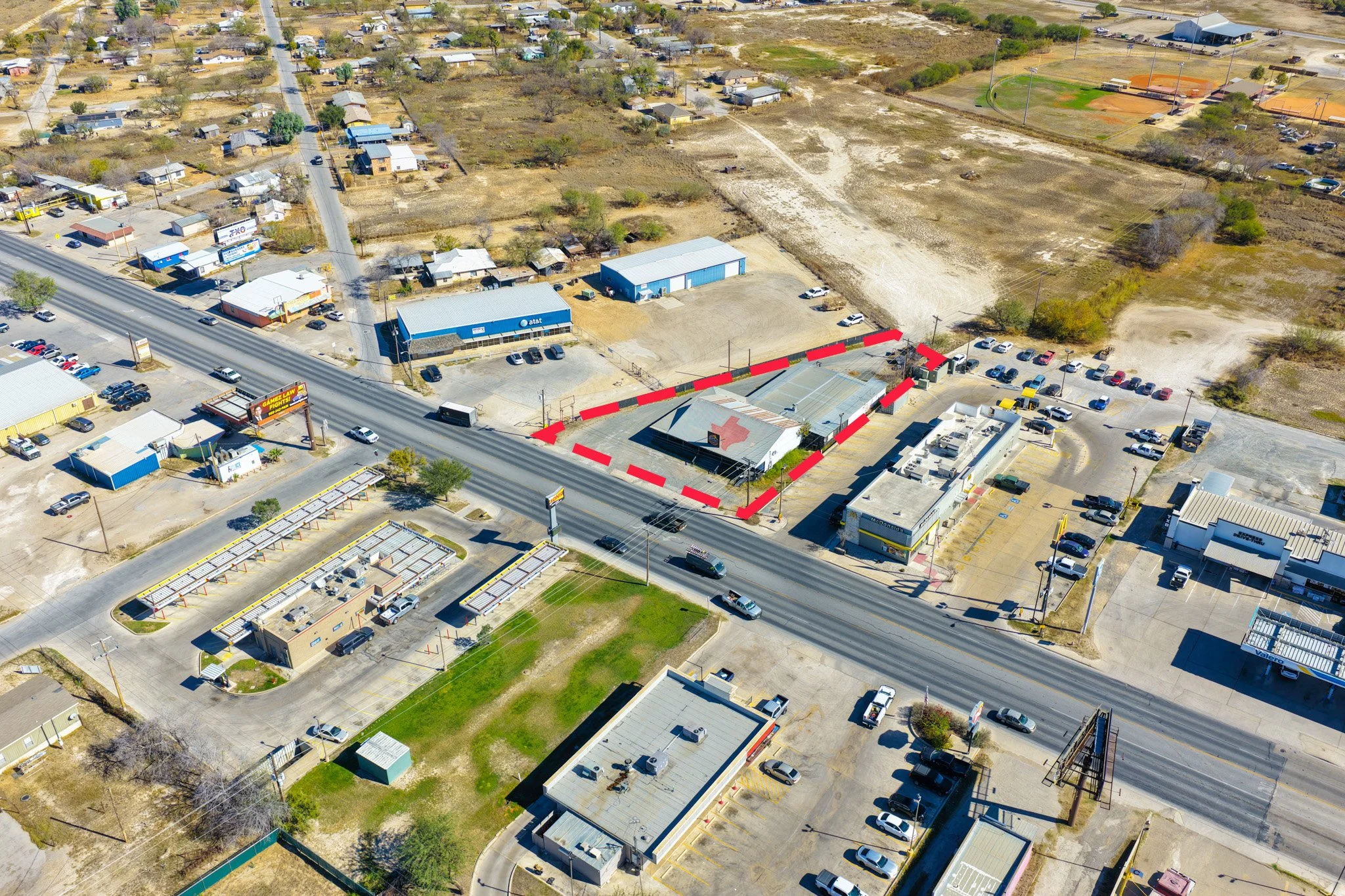 Aerial view of a small commercial area with shops and parking lot along a busy road, with a large open field and residential area in the background.