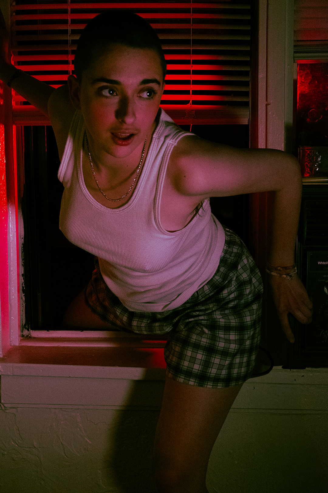 Young woman in a white tank top and plaid shorts crawling out of a window at night with red lighting.