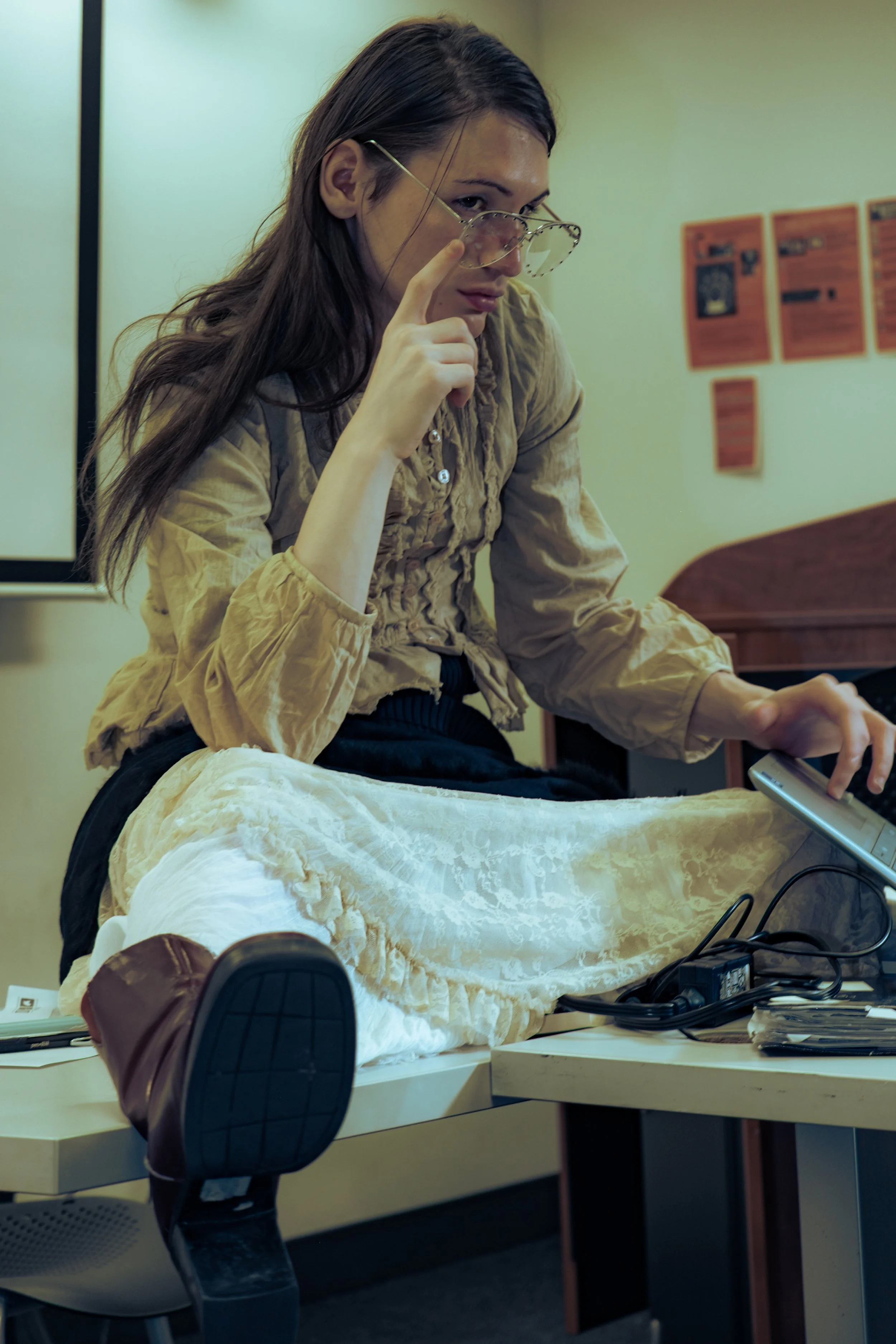 A woman with long brown hair and glasses, wearing a beige blouse, is sitting at a desk, looking over her shoulder at the camera. She appears to be in an office or classroom setting, with a woman in the foreground wearing a white dress and brown shoes. The desk has various electronic devices and cords.
