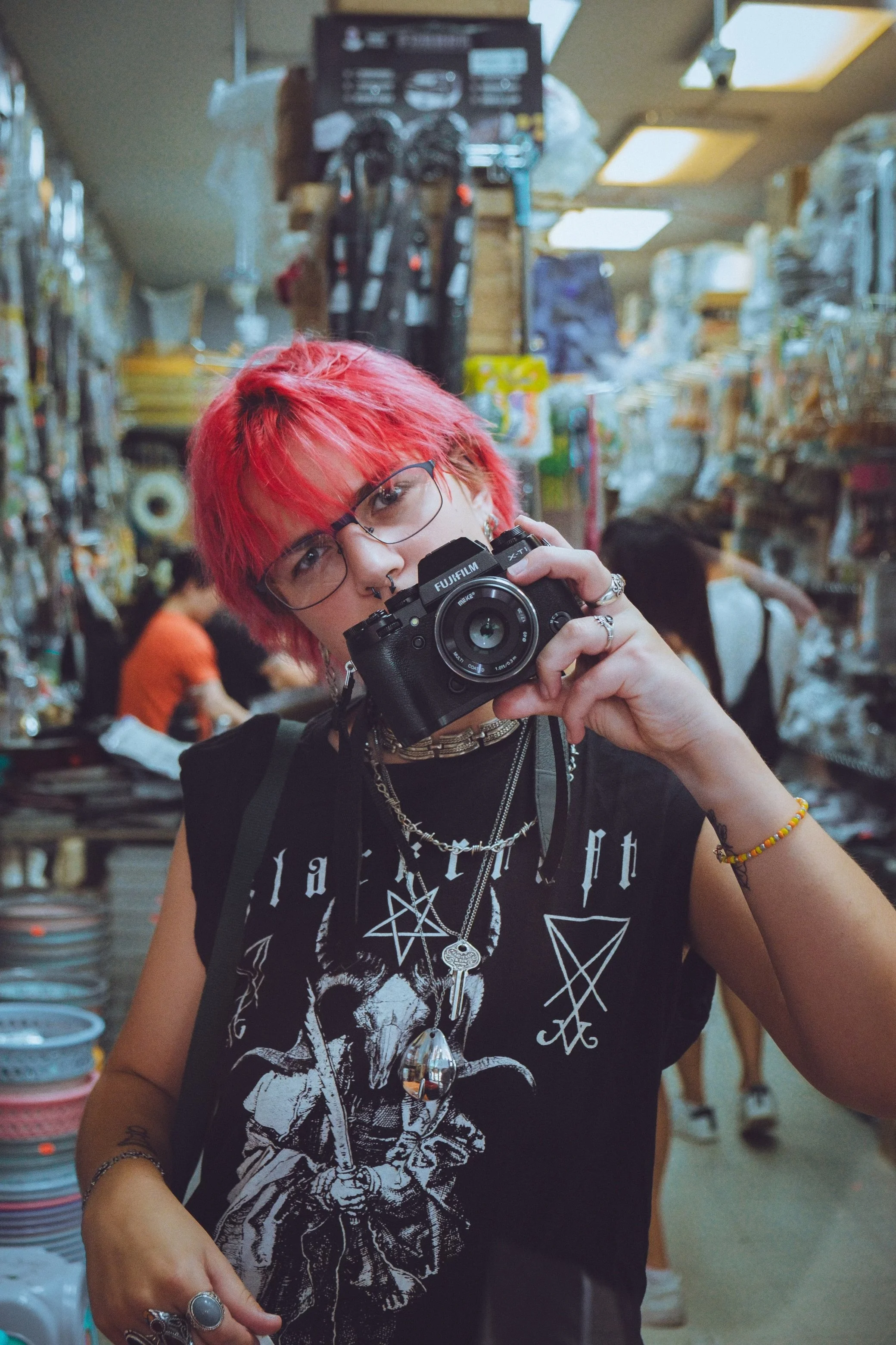 A person with bright red hair and glasses taking a photo with a Fujifilm camera in a store, with shelves of items and other people in the background.