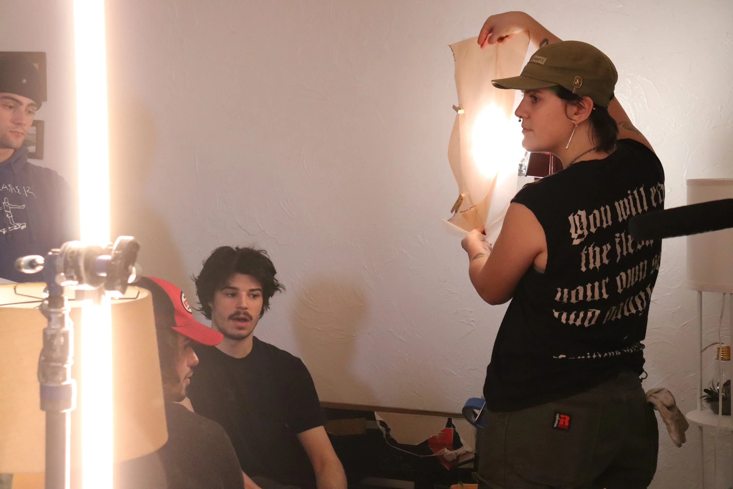 A woman with a cap, earrings, and a septum piercing stands with a group of young men, one wearing a red baseball cap, in a room with a white wall, a lamp, and some decorations.