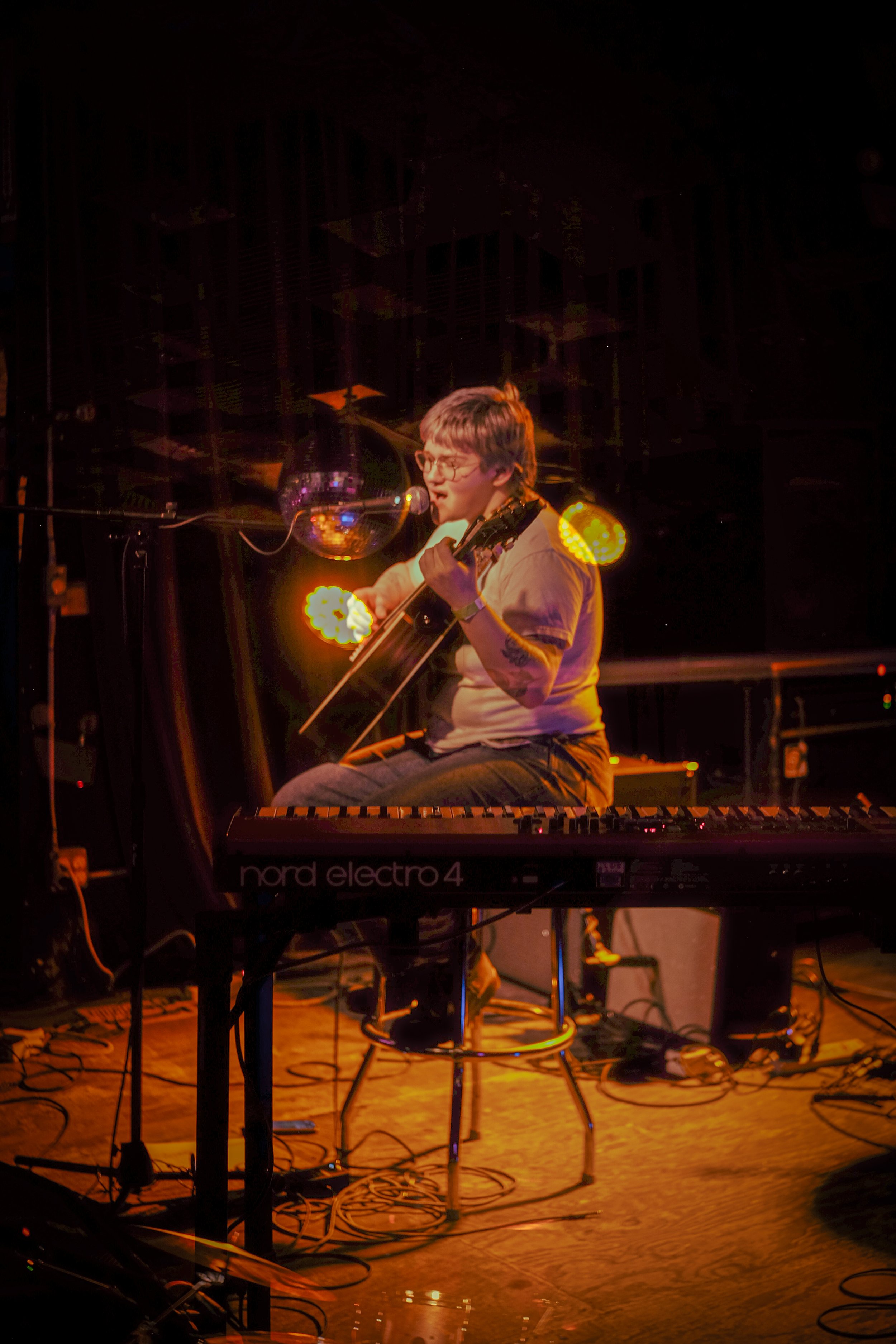Musician playing guitar and singing into a microphone on stage with warm lighting, a keyboard labeled 'Nord Electro 4' in front, and a disco ball reflecting lights.