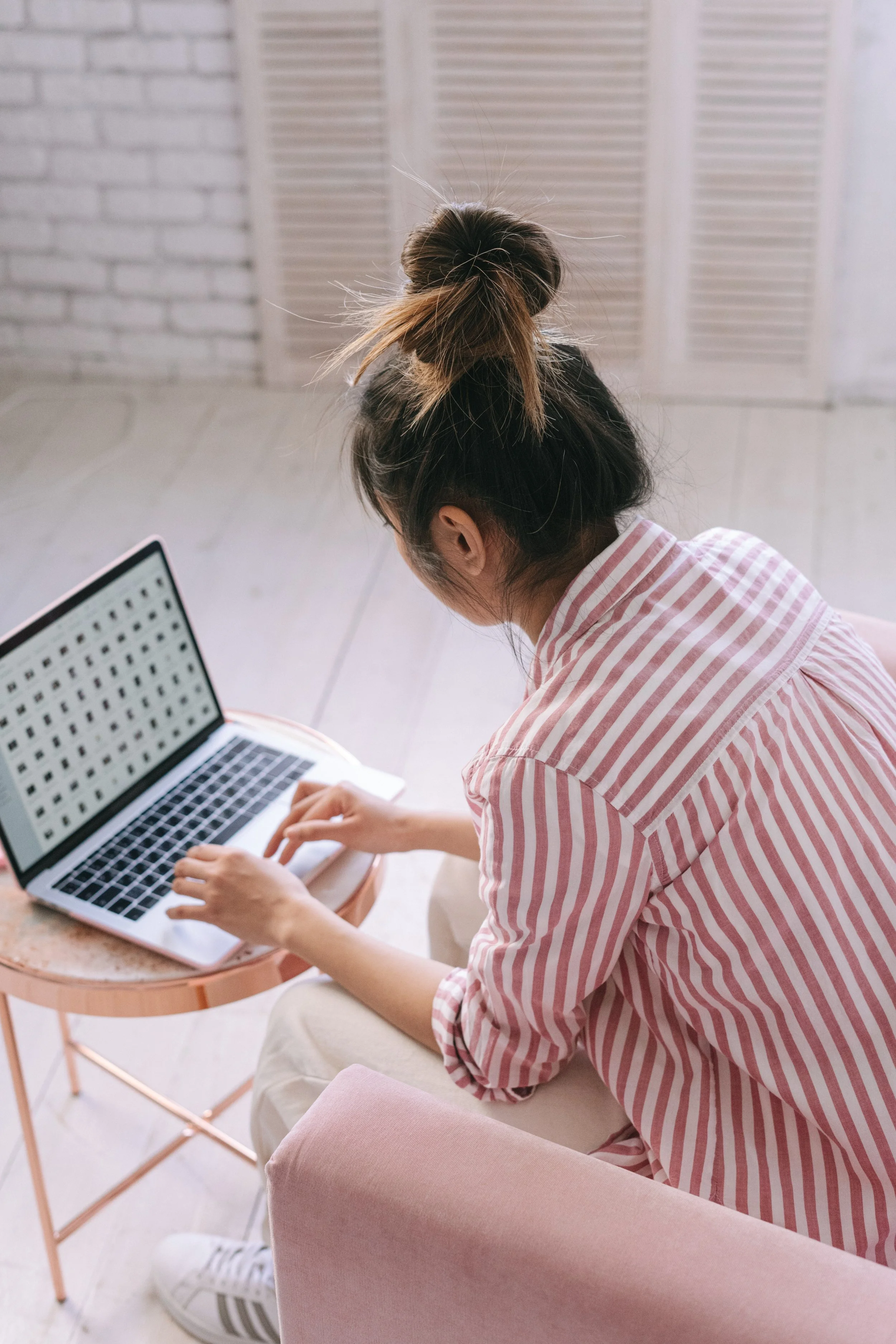 A woman with a messy bun hairstyle, wearing a pink and white striped shirt, sitting at a small round wood table, working on a silver laptop. She is in a room with white brick walls and a white shutter window.