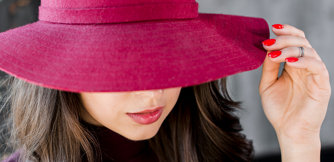 Woman wearing a large pink hat, covering her eyes, with red lipstick and red nail polish, holding the brim of her hat.