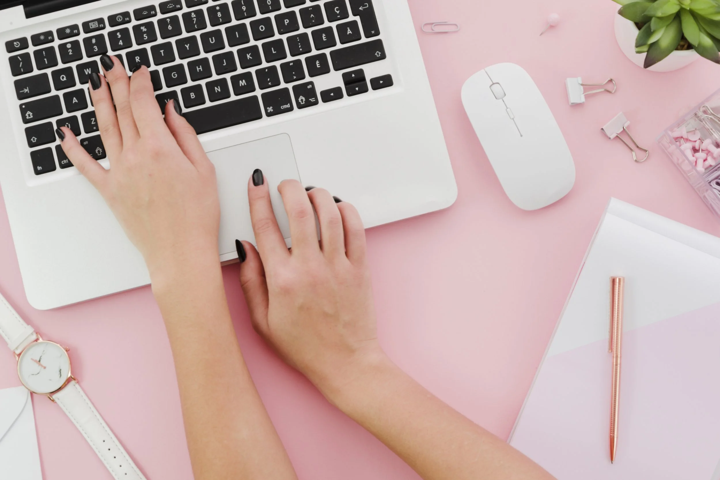 A person using a computer keyboard with black keys on a white laptop, with a white mouse beside it. The workspace is decorated with pink and white office supplies, including a watch, pens, paper clips, a potted plant, and pink and white stationery, all on a pink background.