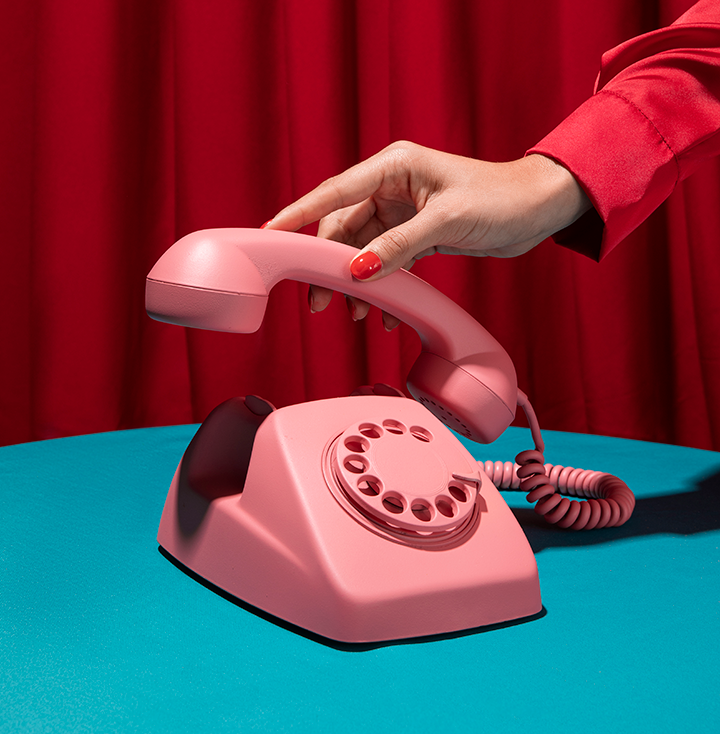 A hand with red painted nails and a red sleeve is picking up a pink rotary telephone, placed on a blue surface with a red curtain in the background.