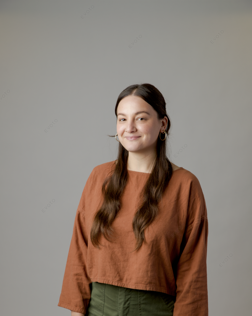 A young woman with long brown hair wearing hoop earrings, a rust-colored long sleeve top, and green pants, smiling softly at the camera against a plain gray background.