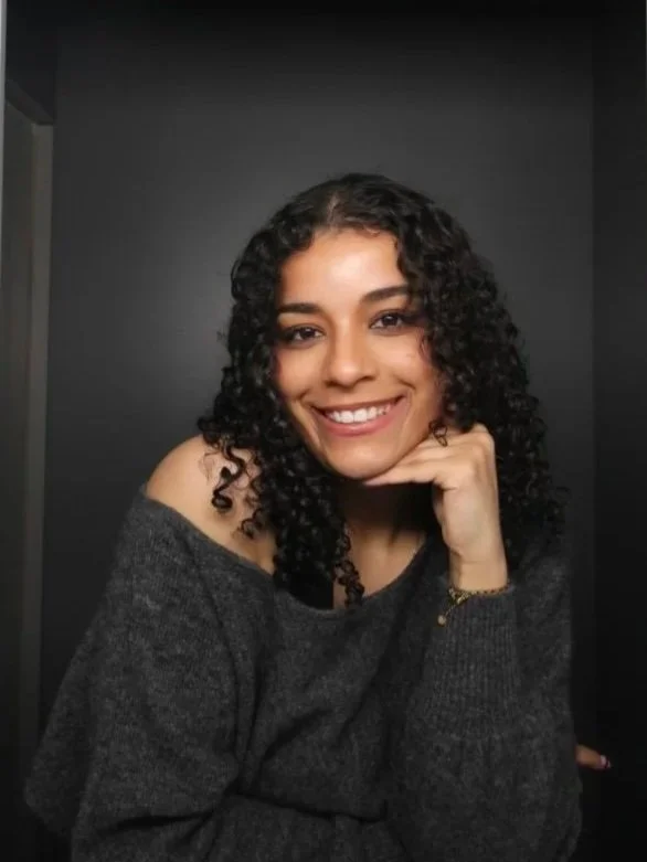 A young woman with curly hair smiling, wearing a dark gray off-the-shoulder sweater, sitting against a black background.