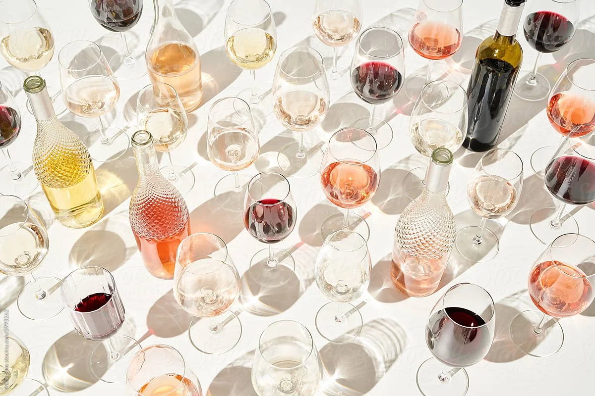 An overhead view of several glasses and bottles of wine and other beverages on a white surface, with sunlight casting shadows.