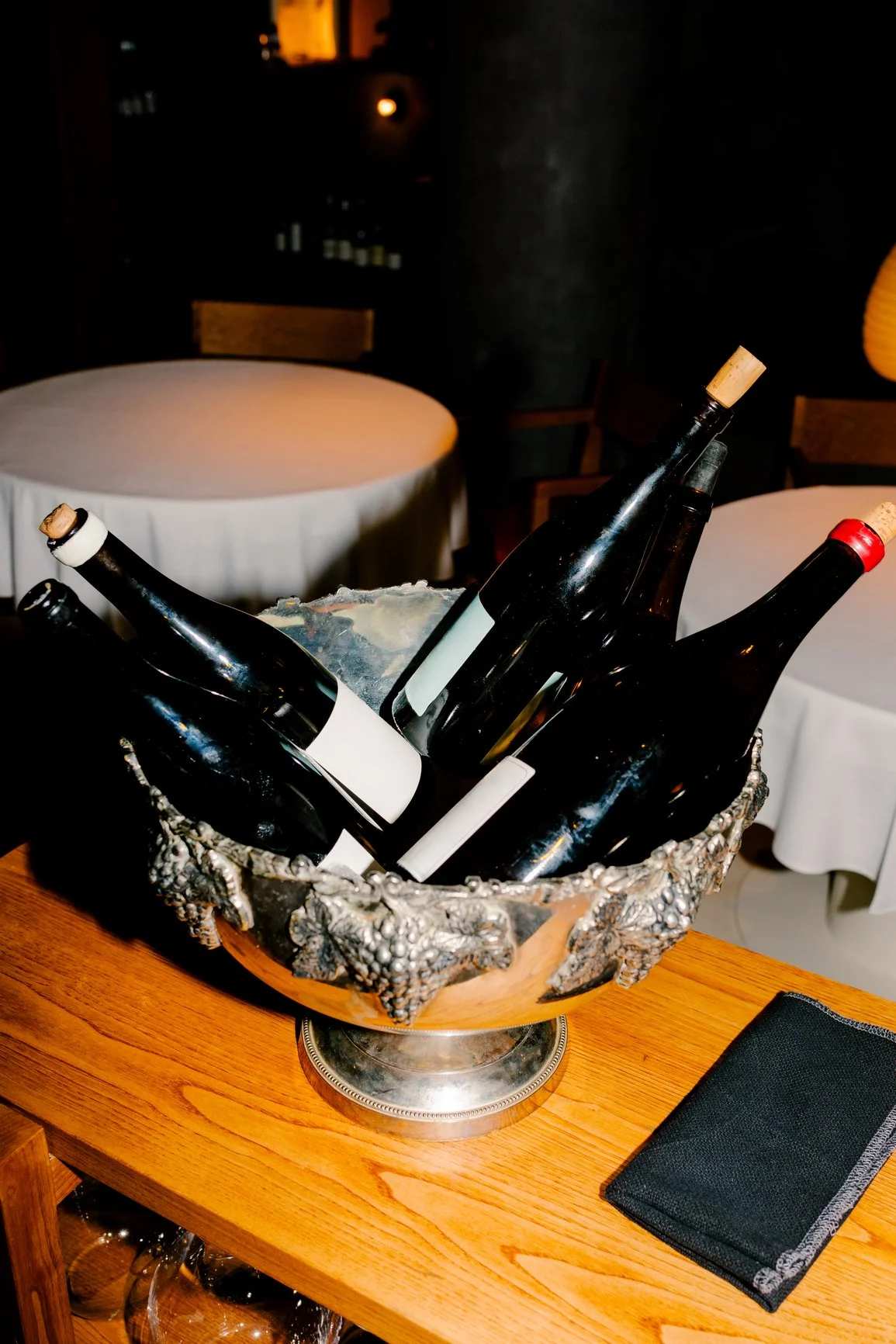 A metal and wood wine bucket filled with ice and empty wine bottles, set on a wooden table in a dimly lit restaurant.