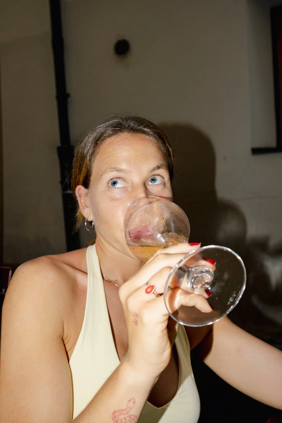 A woman with short brown hair and blue eyes drinking from a large wine glass with white wine. She is wearing a white sleeveless top and red nail polish, with a tattoo of a smiling face on her arm.