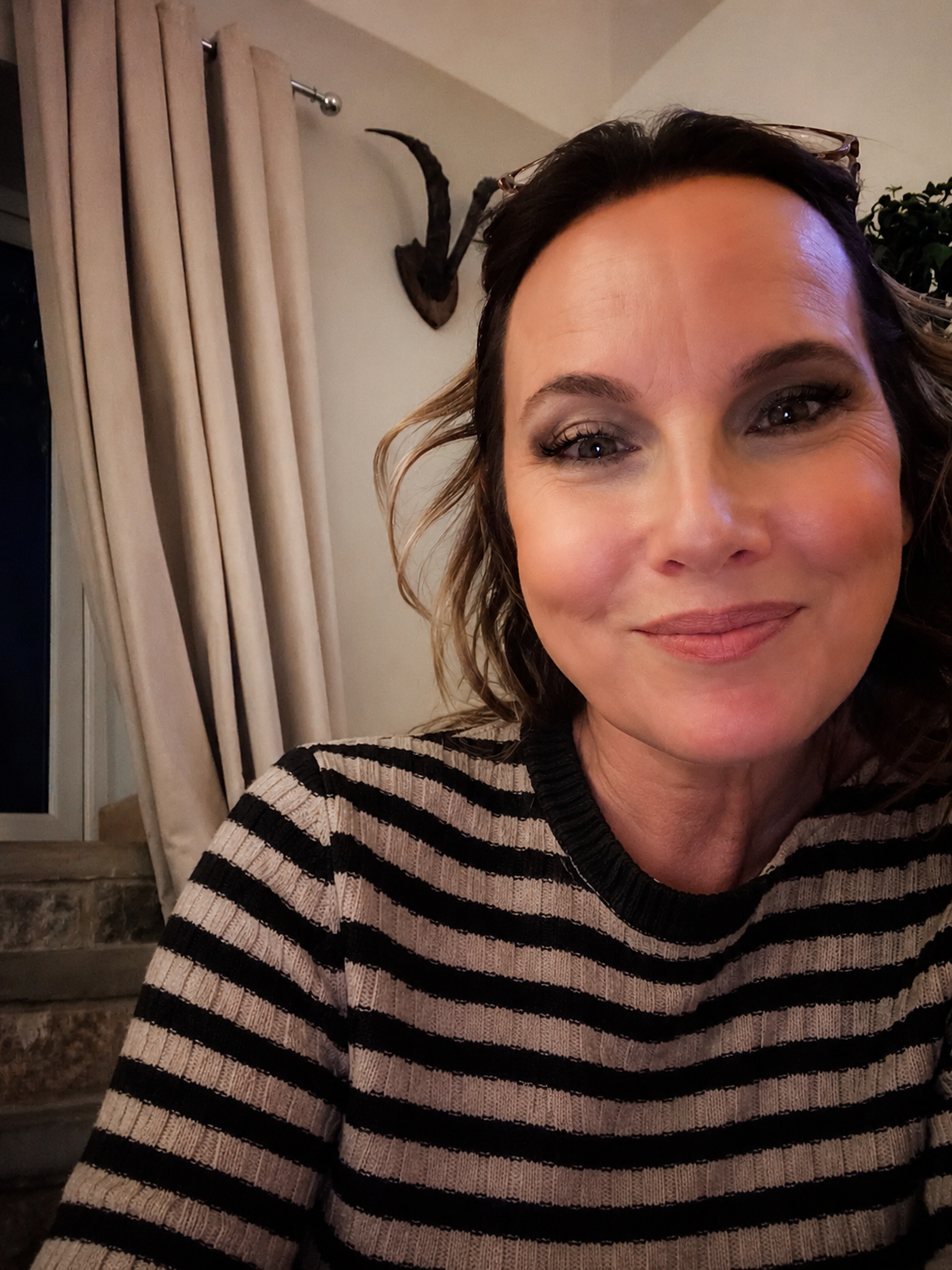Close-up of a woman with brown curly hair, wearing a black and beige striped sweater, smiling in a warmly lit room with beige curtains and a decorative mounted animal skull in the background.
