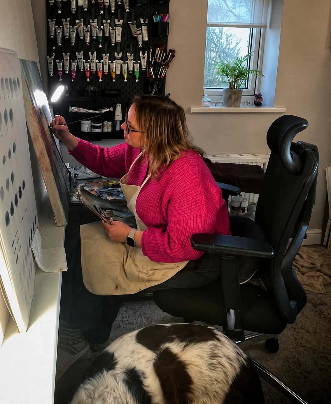 A woman sitting at an art or craft desk, painting or drawing on a canvas with a small brush, wearing a pink sweater and an apron. There is a dog lying on the floor nearby, and art supplies such as paints and brushes on shelves behind her, with a window showing trees outside.