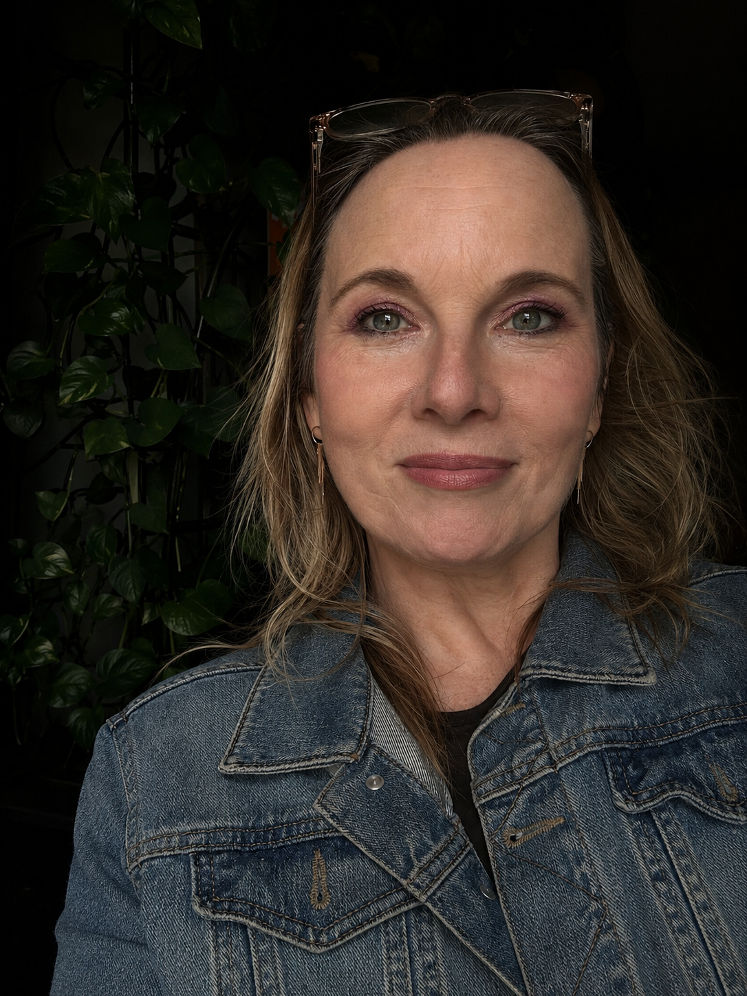 Close-up portrait of a woman with wavy blonde hair, wearing glasses on her head, earrings, and a denim jacket, standing in front of green leafy plants against a dark background.