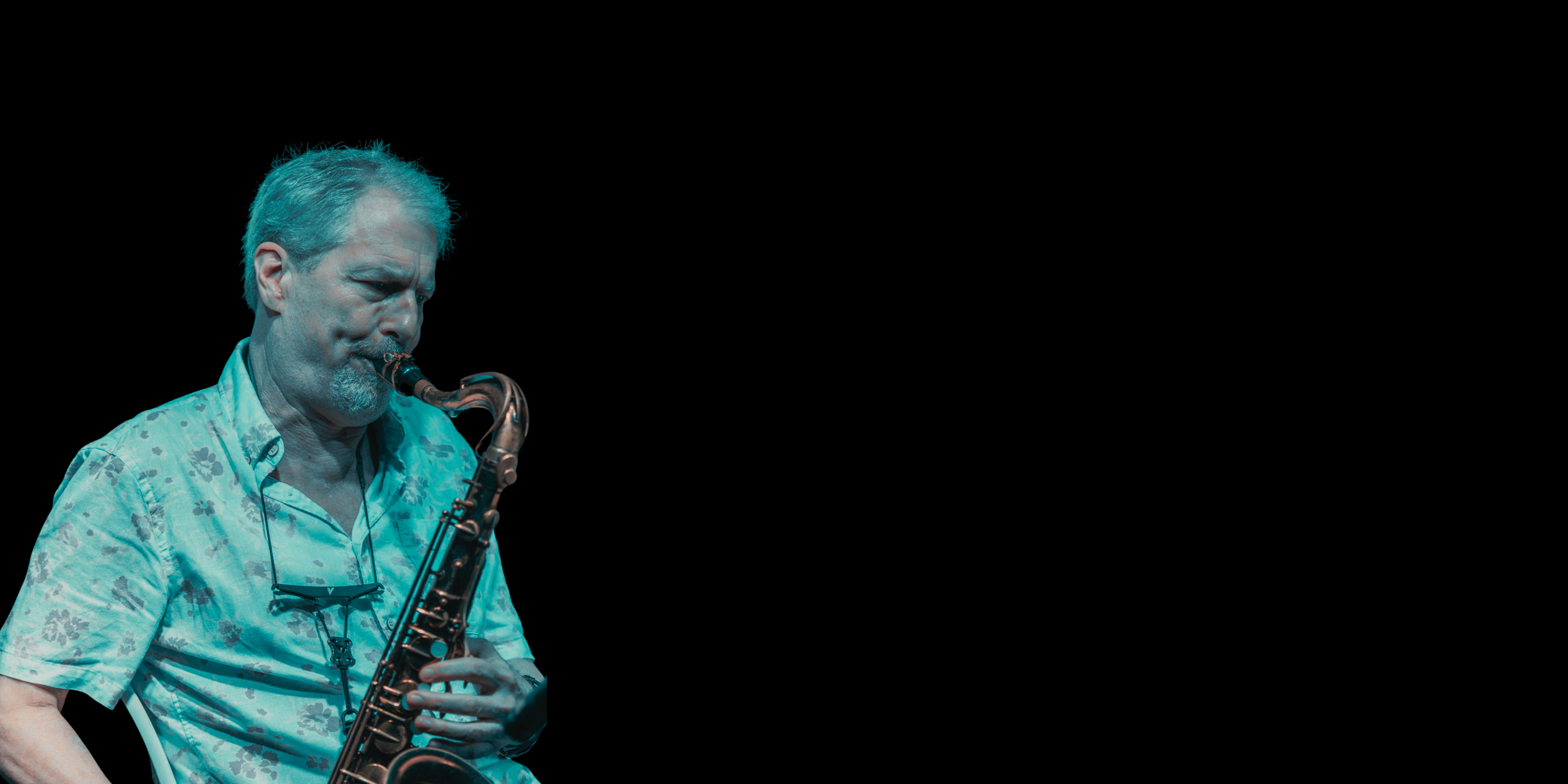 An elderly man with gray hair and a gray beard playing a saxophone against a black background.