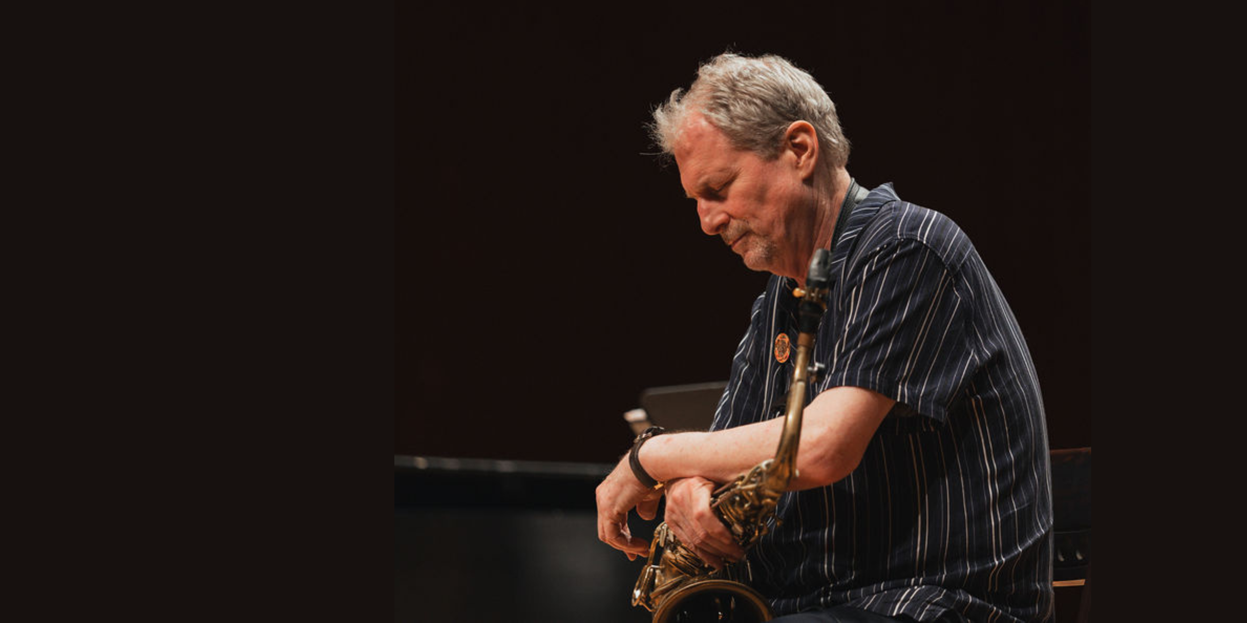 A man with gray hair and a beard, wearing a dark striped shirt, is holding a saxophone and looking down thoughtfully against a dark background.
