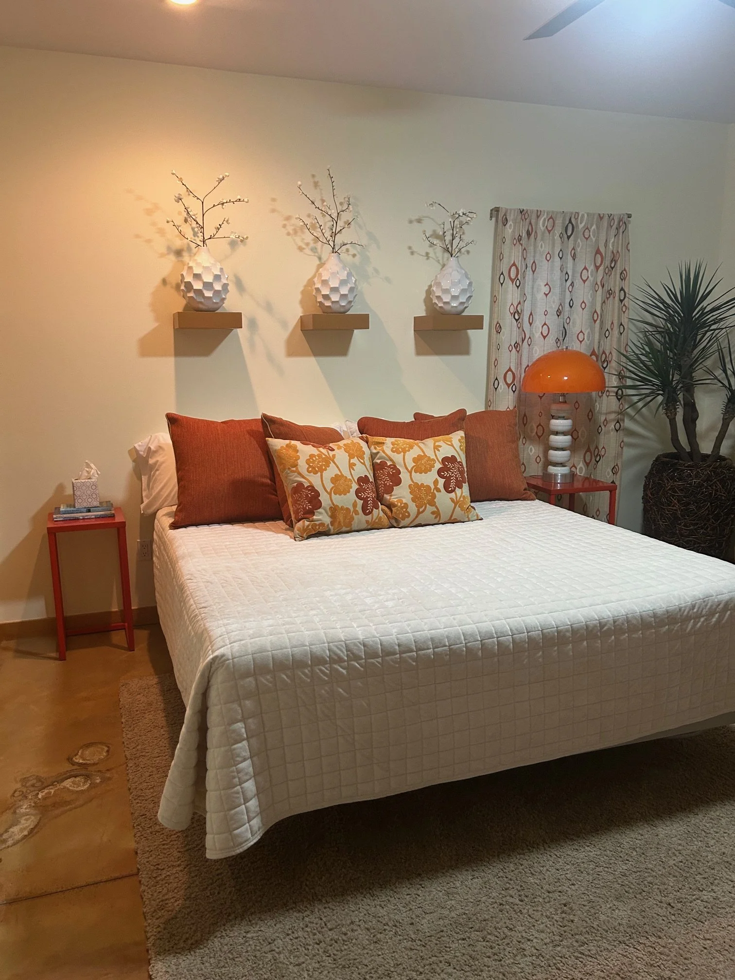 A bedroom with a bed covered in a white quilt, decorated with multiple orange, brown, and white patterned pillows. To the right of the bed is a red side table with books and a tissue box. A large pot with a tall plant is in the corner. On the wall ab