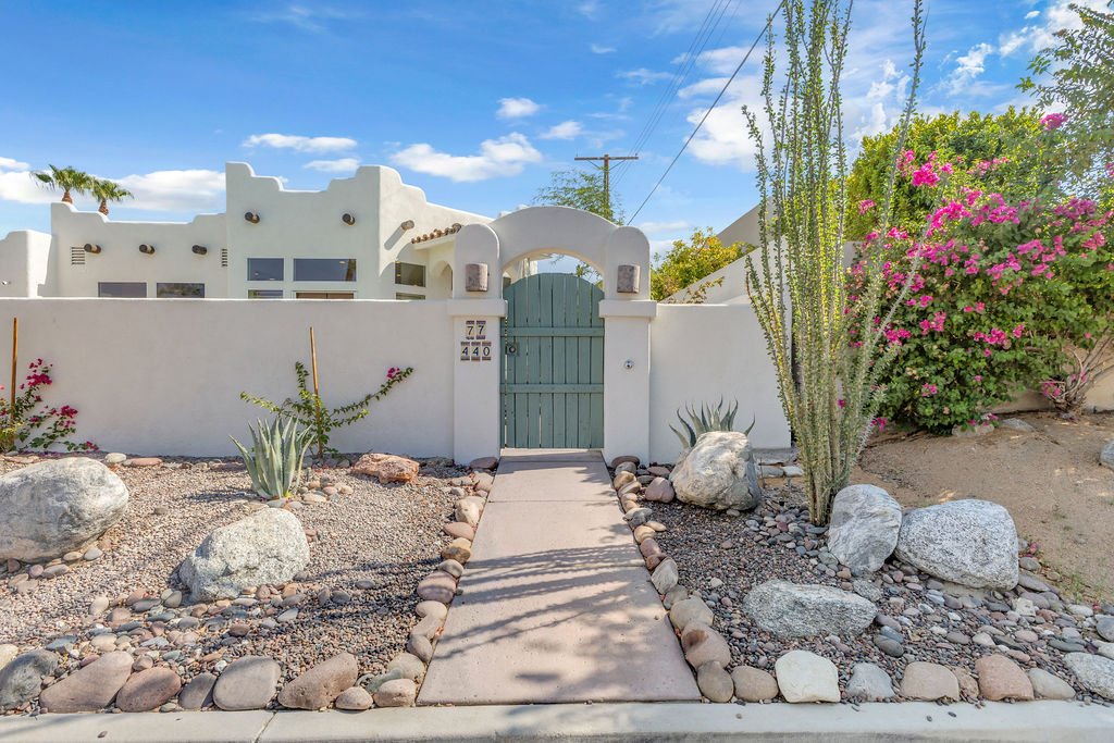 A white stucco wall with a wooden gate, desert landscaping with rocks and cacti, and pink bougainvillea bushes in the front yard under a blue sky with some clouds.