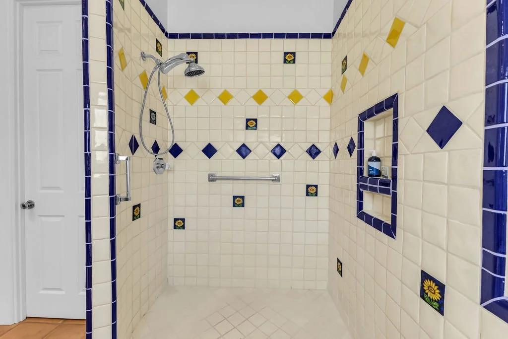 A walk-in shower with cream-colored tiles, decorated with blue and yellow accent tiles featuring sunflowers, and a built-in shelf with a bottle of body wash.