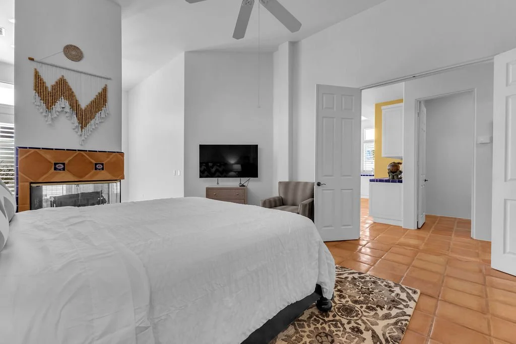 A bedroom with a white bed, wall art, a fireplace, a wall-mounted TV, an armchair, and an open doorway leading to a kitchen with yellow and white cabinets.