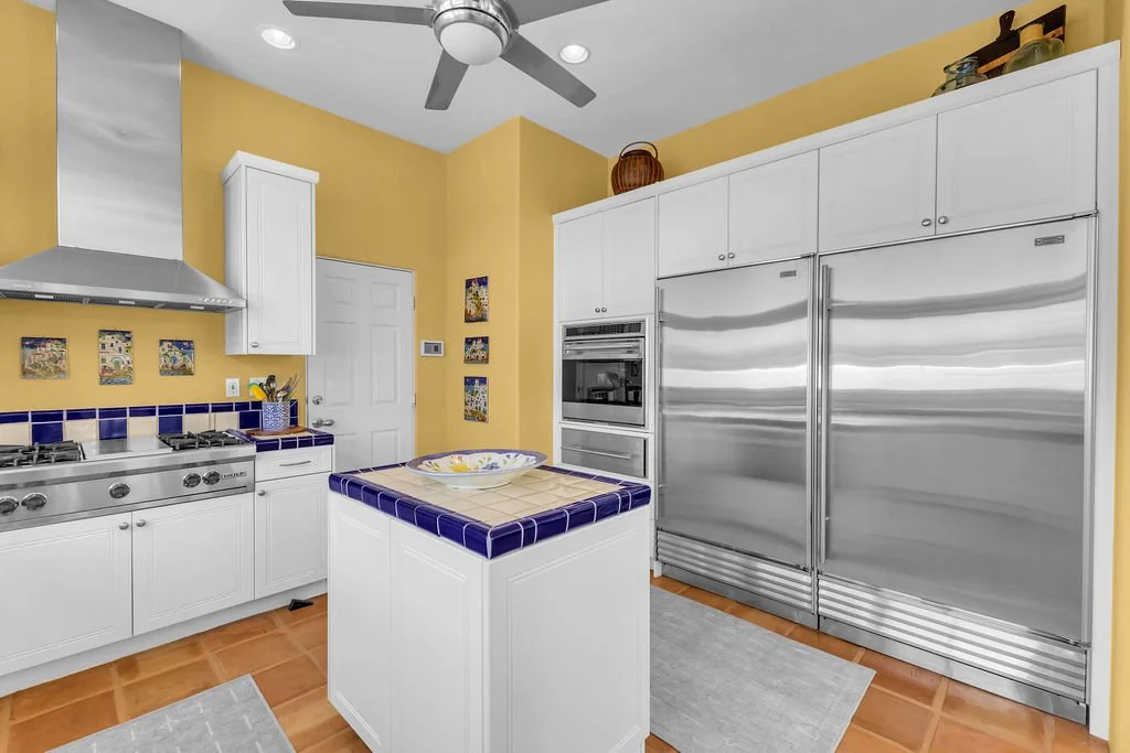 Kitchen with yellow walls, white cabinets, a stainless steel refrigerator, double oven, gas stove, and a small island with a blue and beige tiled countertop and a decorative bowl.