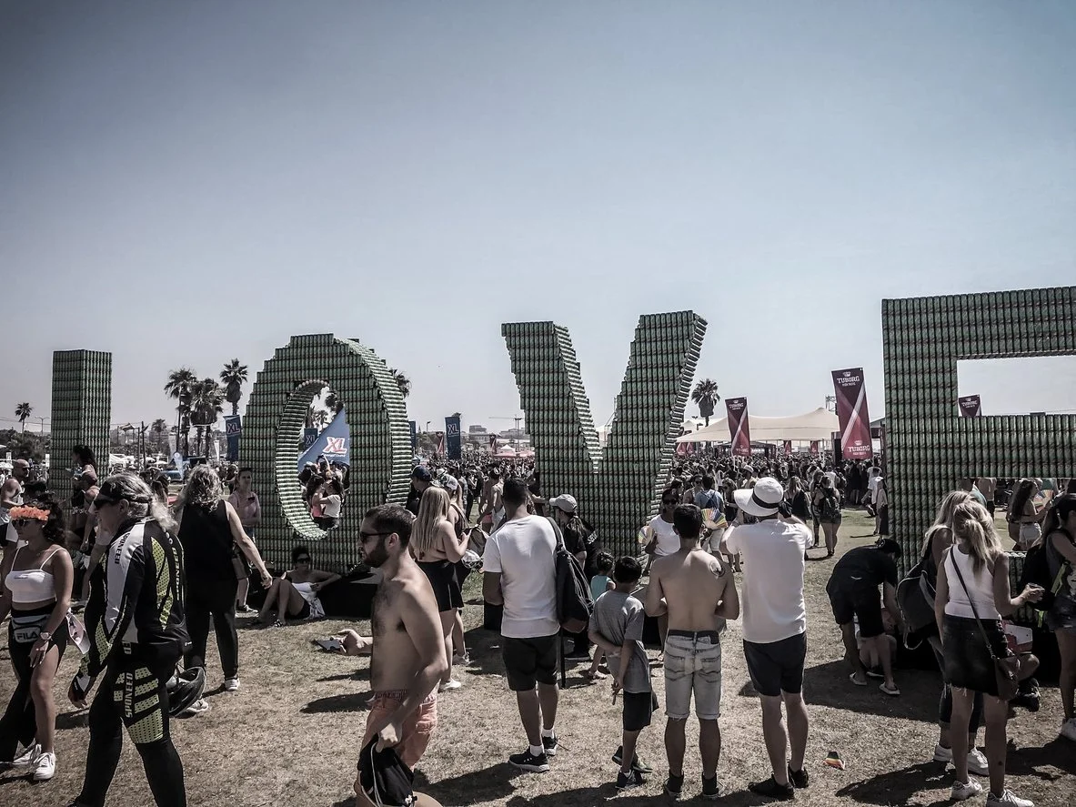 Large outdoor festival with a crowd of people, some dressed casually and others in summer attire. There are three big block letters spelling 'LOVE' made out of green bottles or cans in the background, along with palm trees and tents.