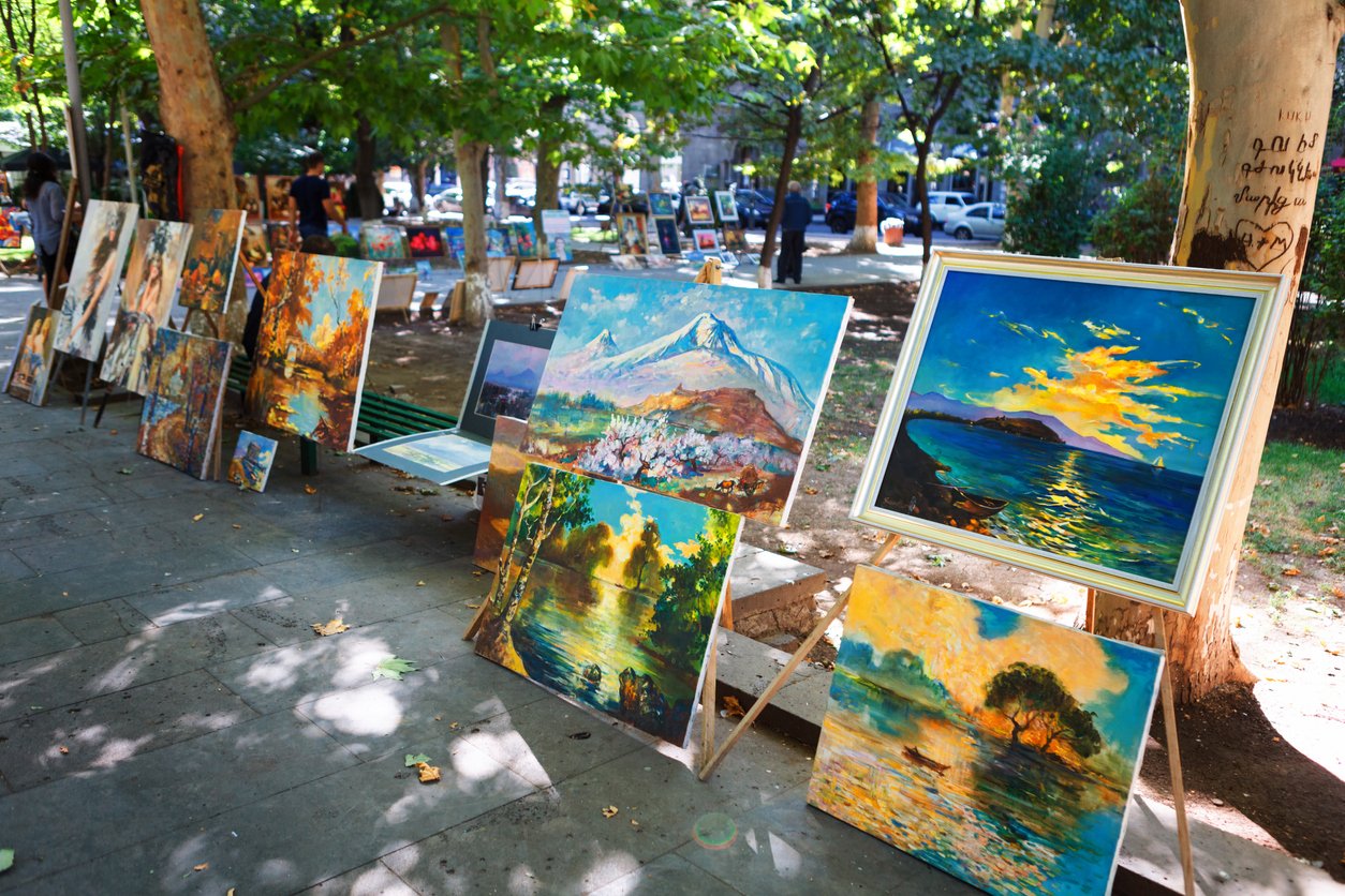 Outdoor art gallery displaying numerous colorful landscape and nature paintings along a city sidewalk shaded by trees.