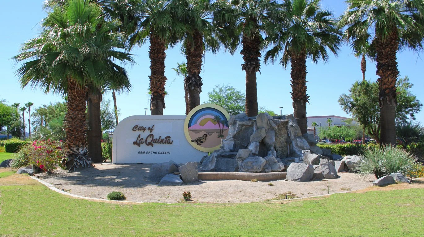 City of La Quinta sign with palm trees and a water fountain in front of it, located in a landscaped area with grass and plants.