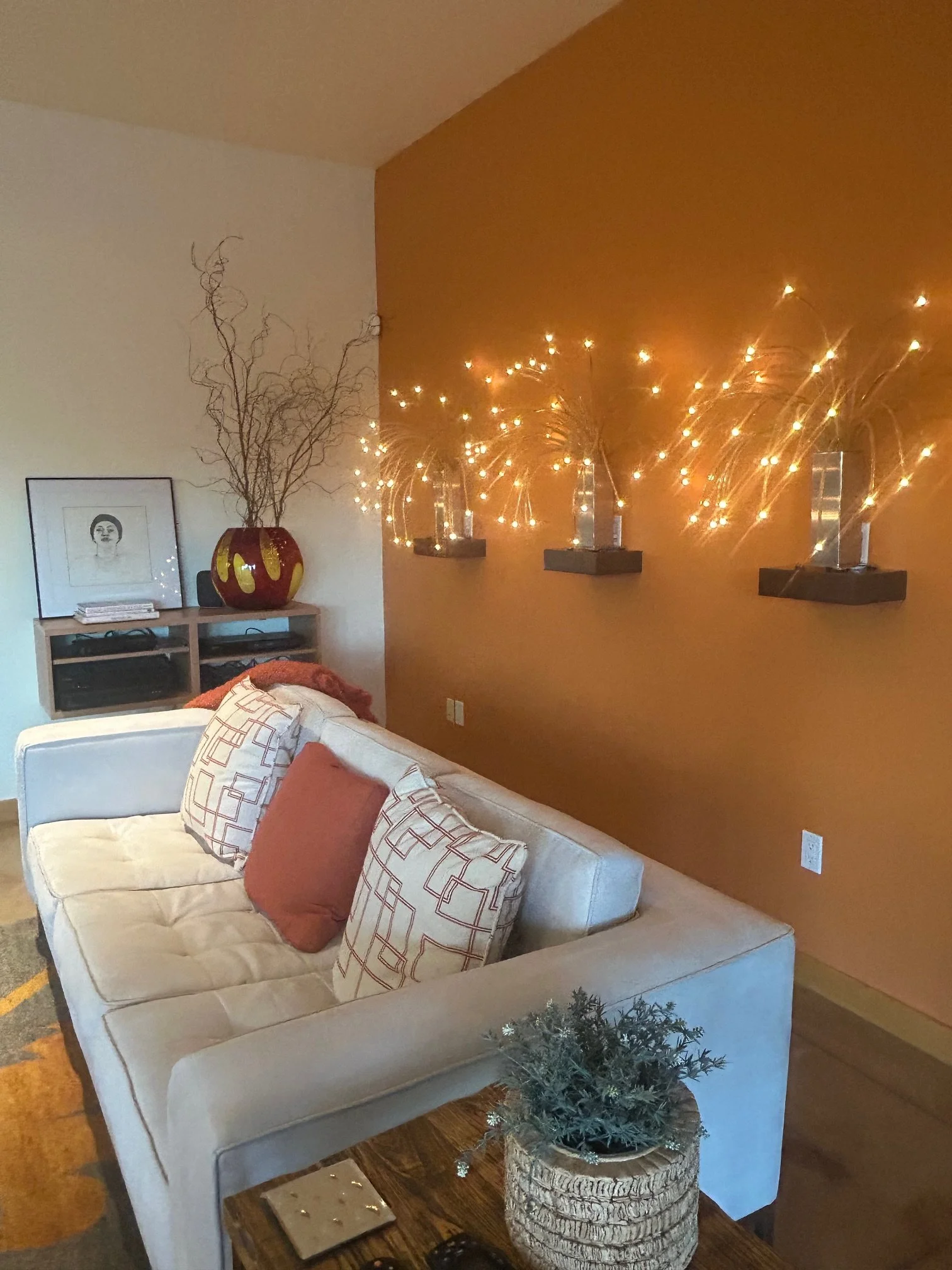 Living room with beige love seat, decorative pillows, a small table with a small artificial Christmas tree in a textured pot, a wall with string lights and shelves, and artwork on the wall.