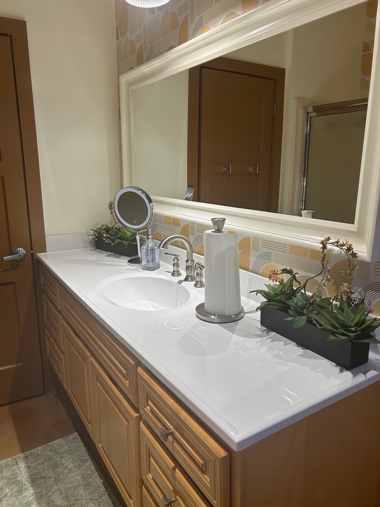 Bathroom mirror reflecting a door, a vanity with plants, soap, paper towel roll, and a faucet. The wall behind has geometric patterned wallpaper.