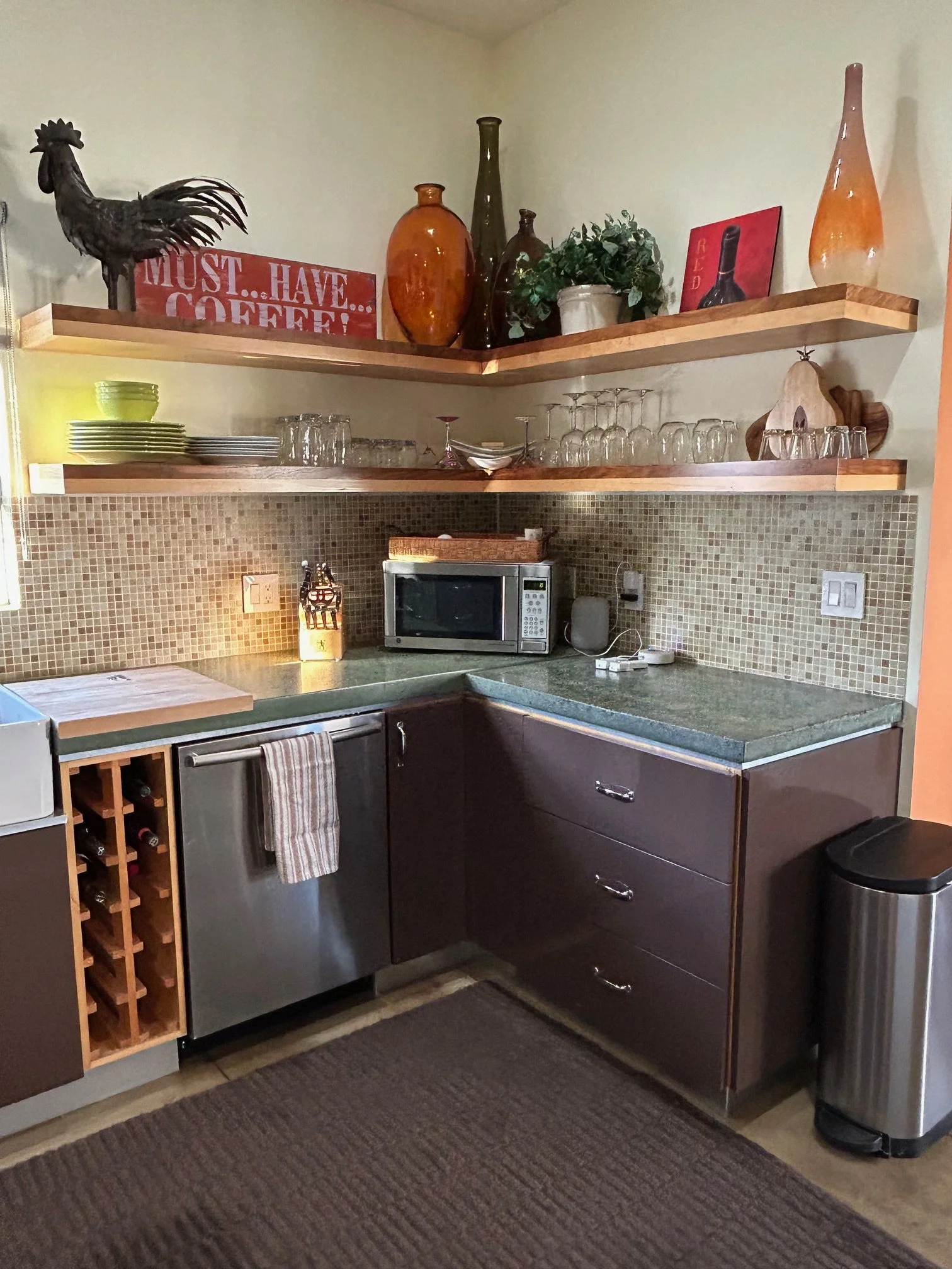 A kitchen corner with two floating wooden shelves holding decorative items, dishes, and glasses. Below the shelves is a countertop with a microwave, a small wine rack, and a trash can. The wall has a small mosaic tile backsplash.