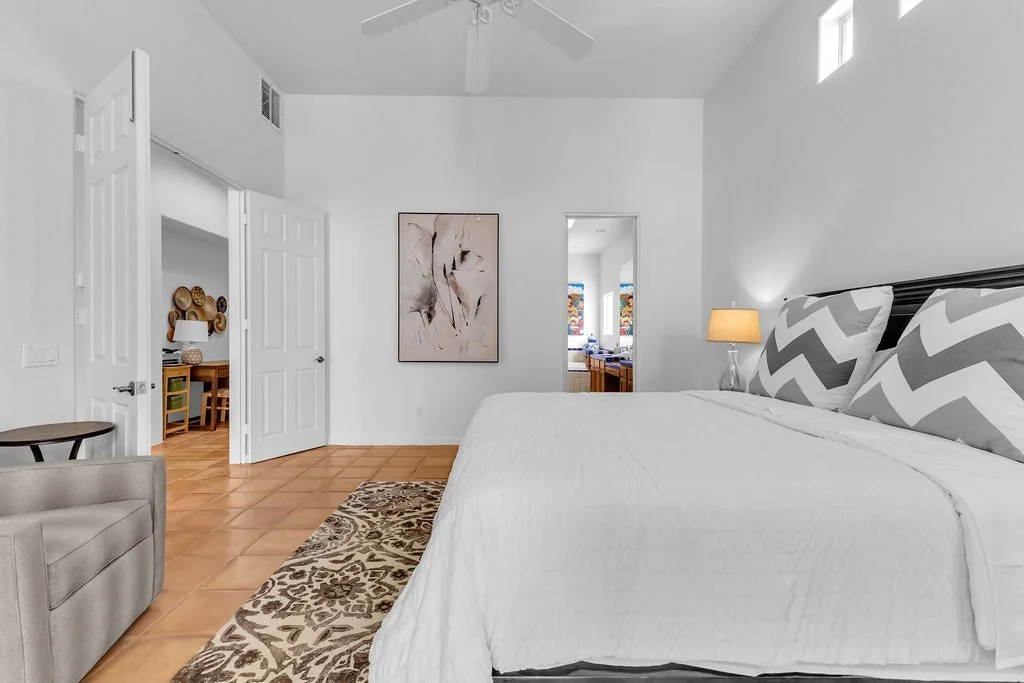 A bedroom with white walls, a large bed with gray and white chevron pillows, a bedside lamp, a ceiling fan, and two small high windows. An abstract painting hangs on the wall, and an open door reveals a hallway with a dining area.