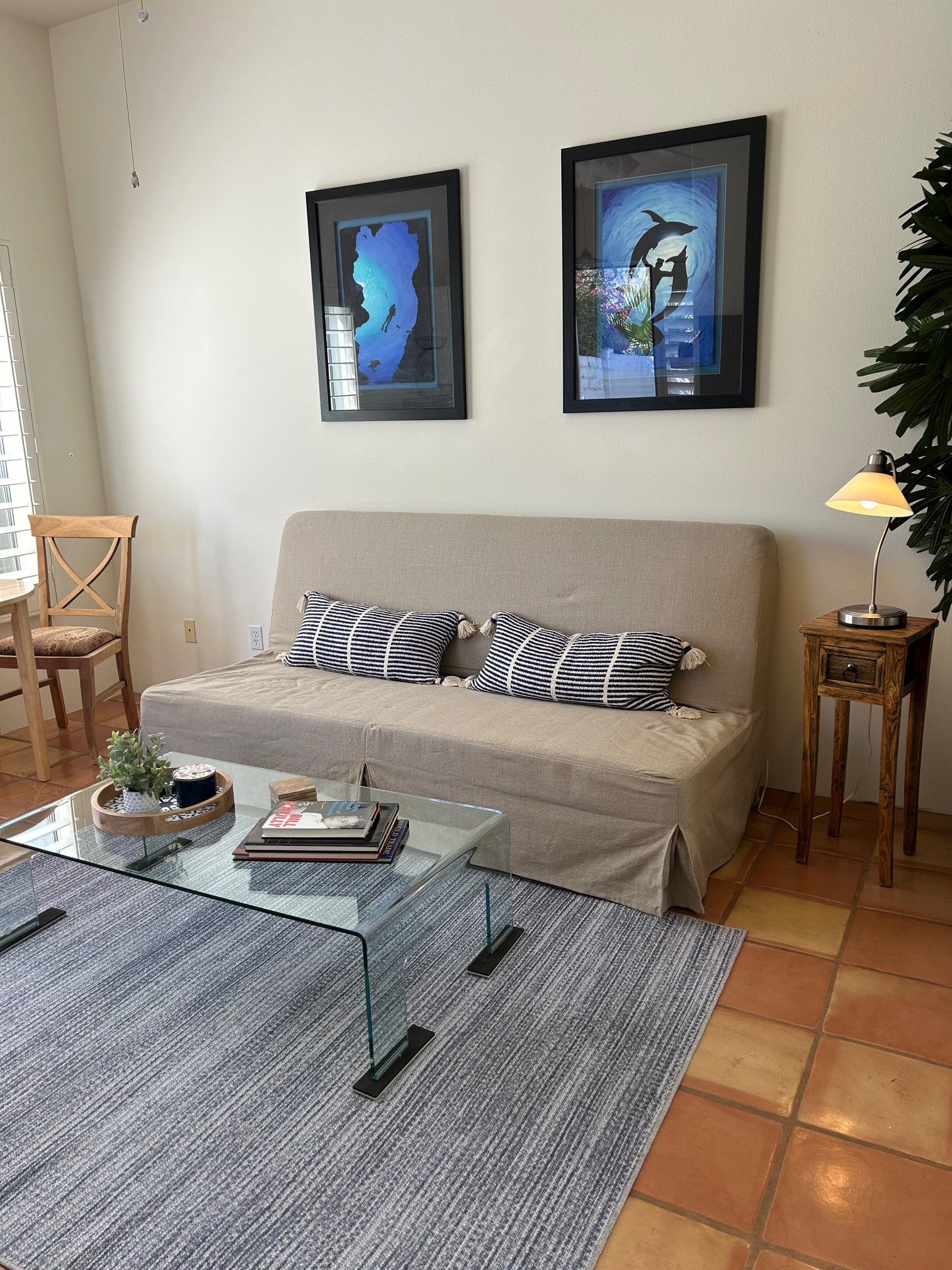 Living room with beige sofa adorned with striped pillows, a glass coffee table with magazines and a tray, a small wooden side table with a lamp, two framed nautical-themed pictures on the wall, a potted plant, and a window with white blinds.
