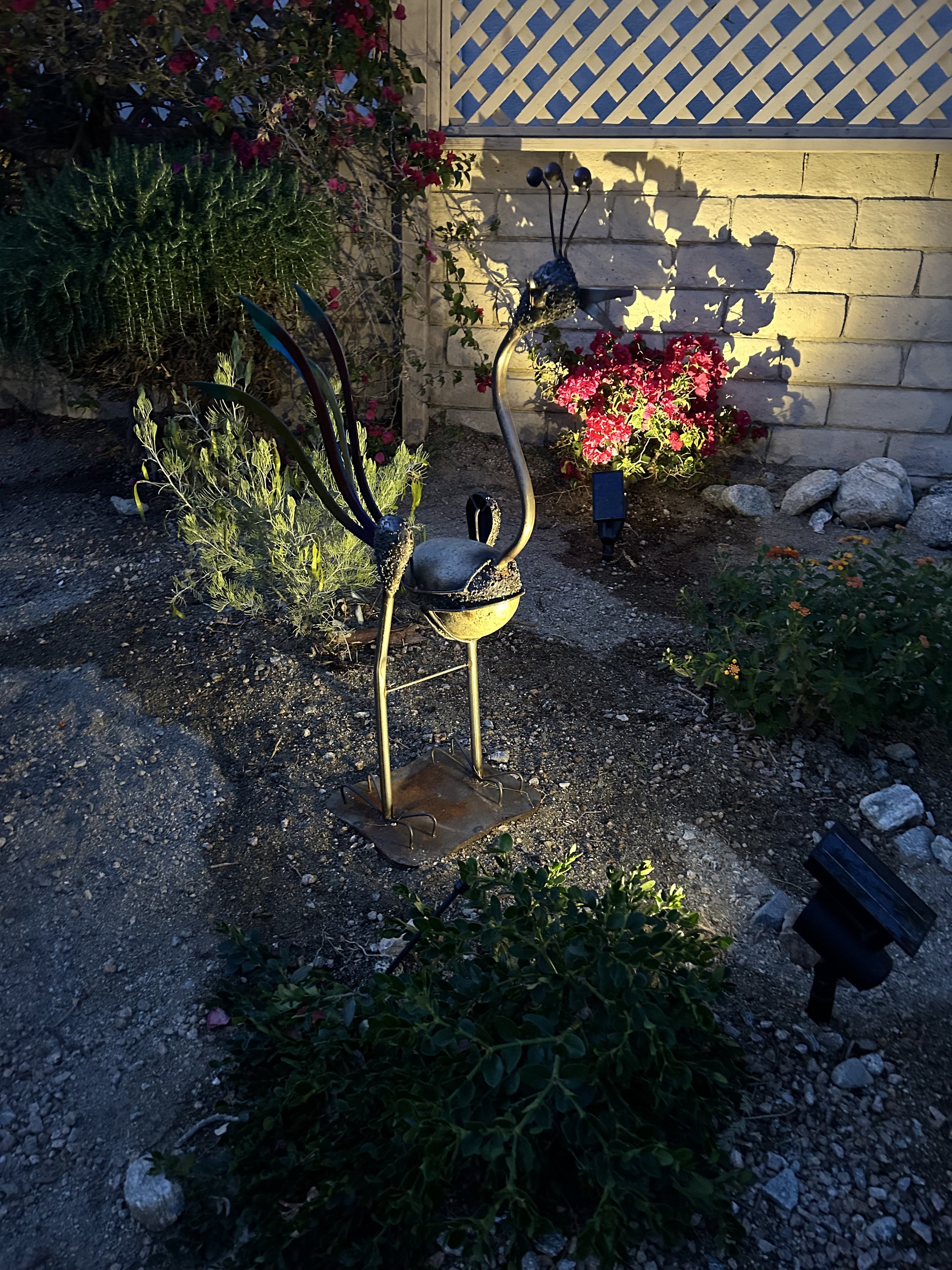 Decorative garden sculpture of a peacock with colorful tail feathers, placed in front of flowering plants and a brick wall with lattice fencing, illuminated by a garden light at dusk.