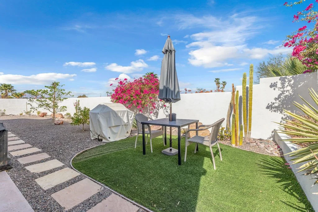 A backyard patio with an artificial grass patch, outdoor table, chairs, and a closed umbrella. There are desert plants including cacti and pink flowering bushes, with a white wall and a bright blue sky in the background.
