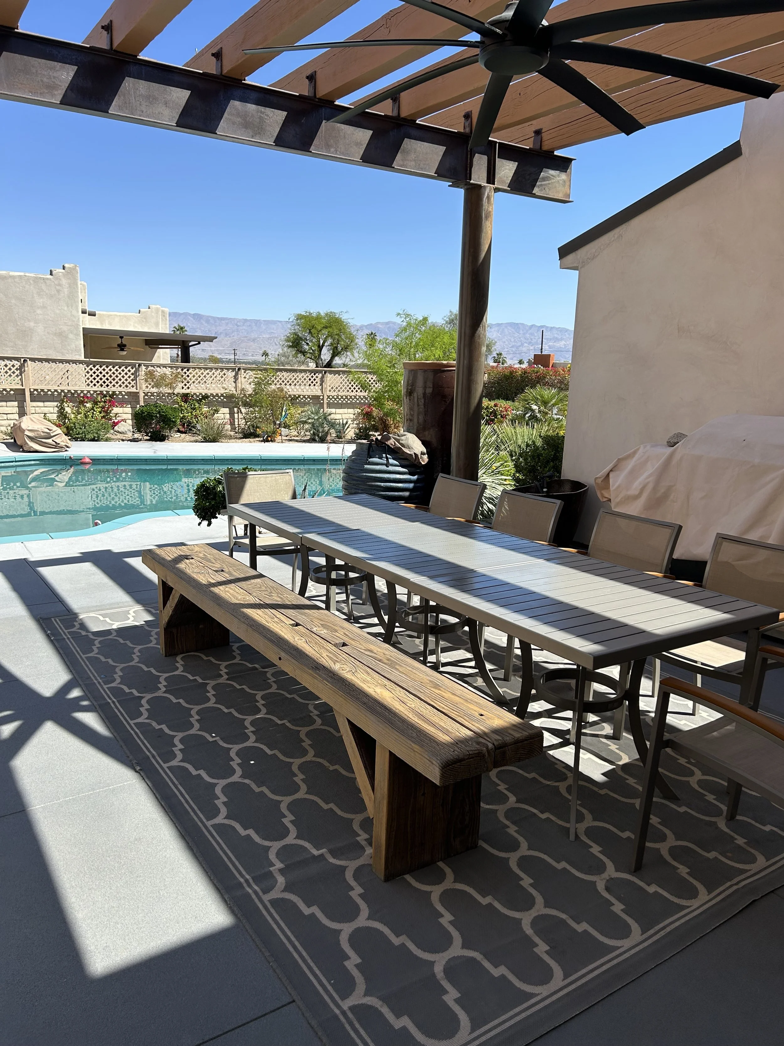 Patio area with a dining table and chairs, a rug, a swimming pool, a shaded sitting area with a cover, a large pot with plants, and desert mountain landscape in the background.