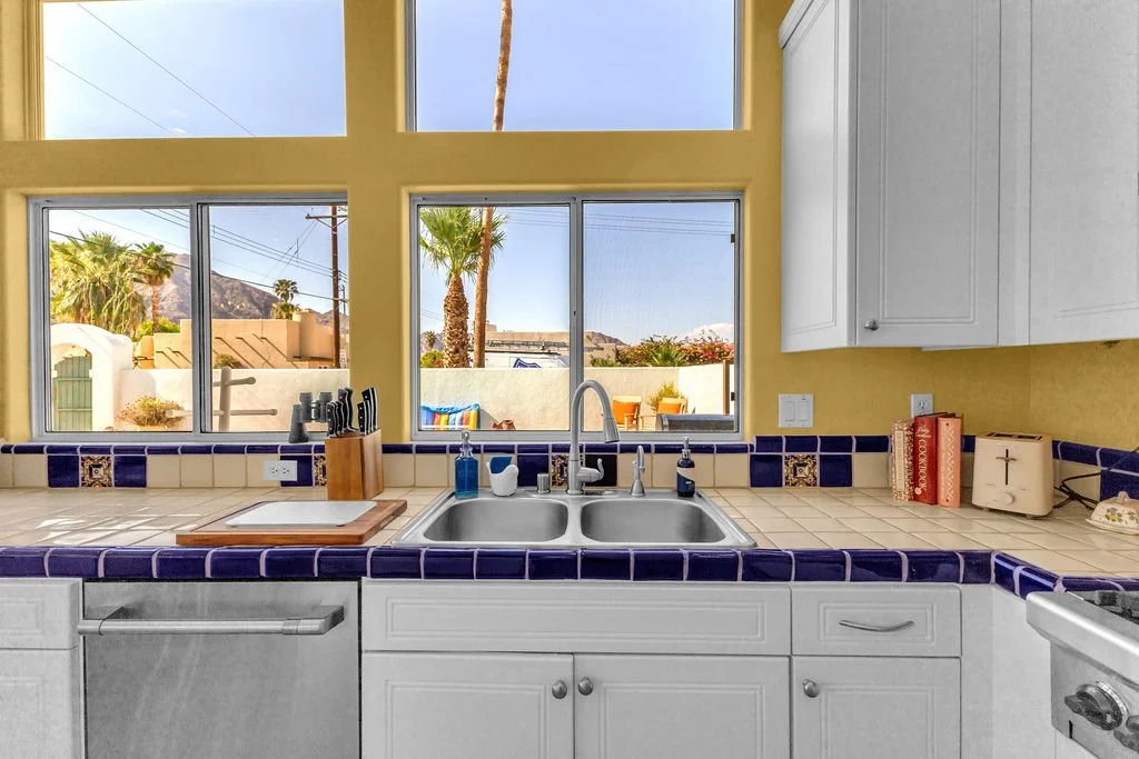 View from kitchen sink showing large windows with a backyard with palm trees, outdoor chairs, and a mountain in the background.