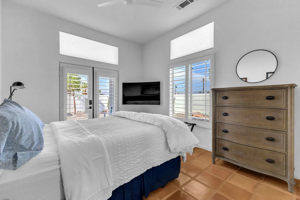 A bright bedroom with a white bed, a wooden dresser, a wall-mounted TV, and large windows with white shutters.