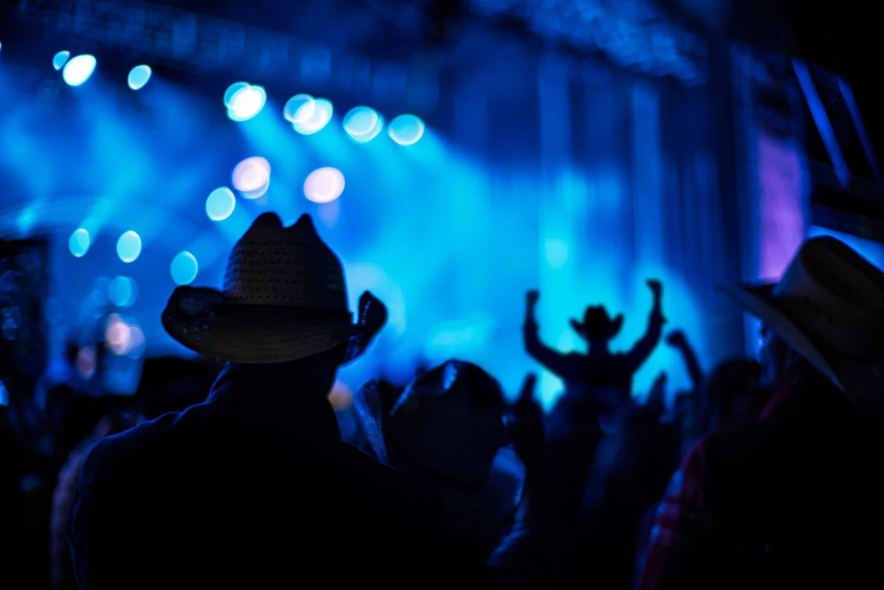 People wearing cowboy hats attending a concert with blue stage lights and a performer in the background raising their arms.