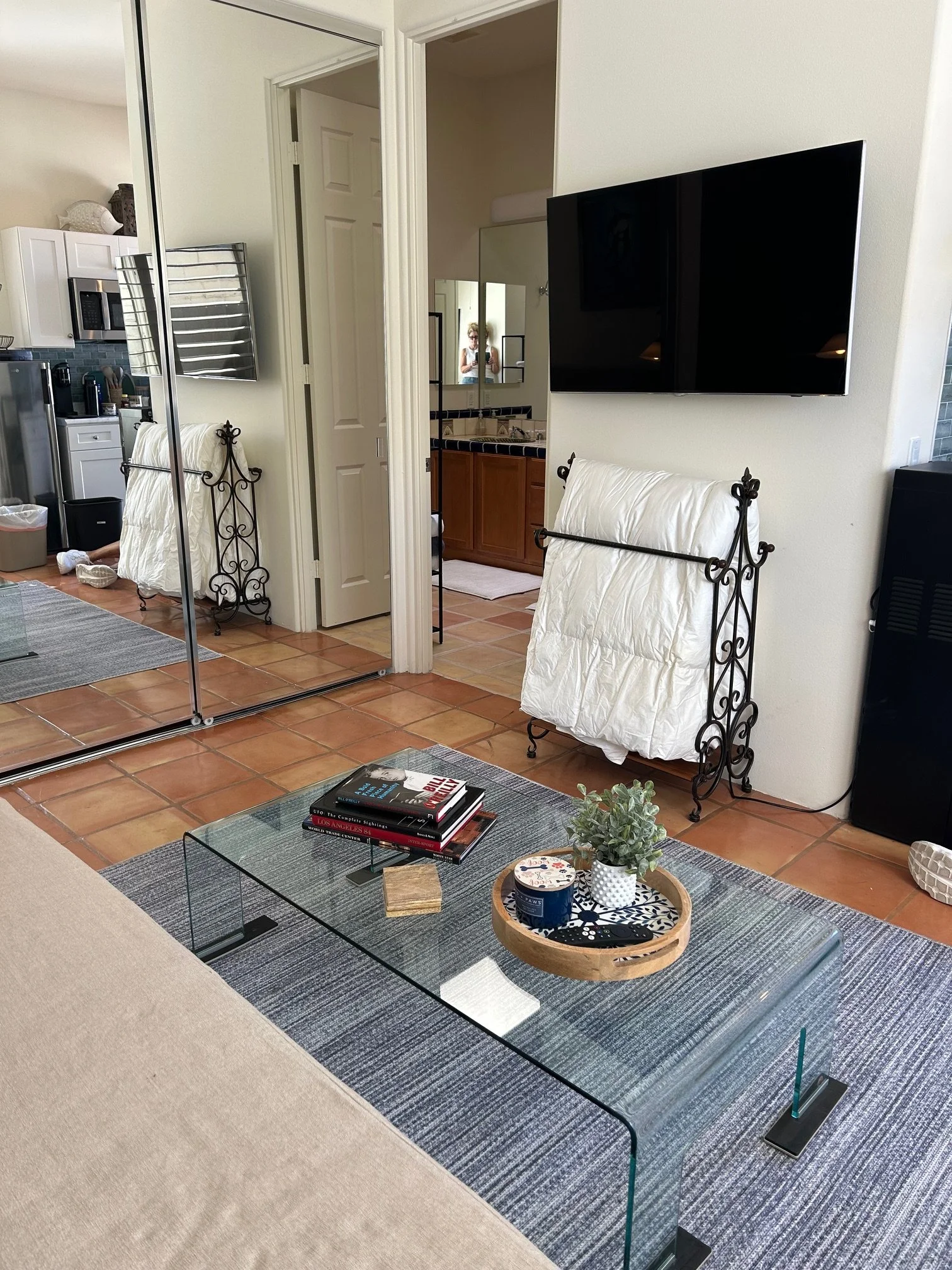 Living room with a glass coffee table holding books, a small plant, and a decorative tray, with a beige sofa in the foreground, a blue rug, and a wall-mounted TV. Behind the coffee table, there are two white covered chairs with decorative metal frame
