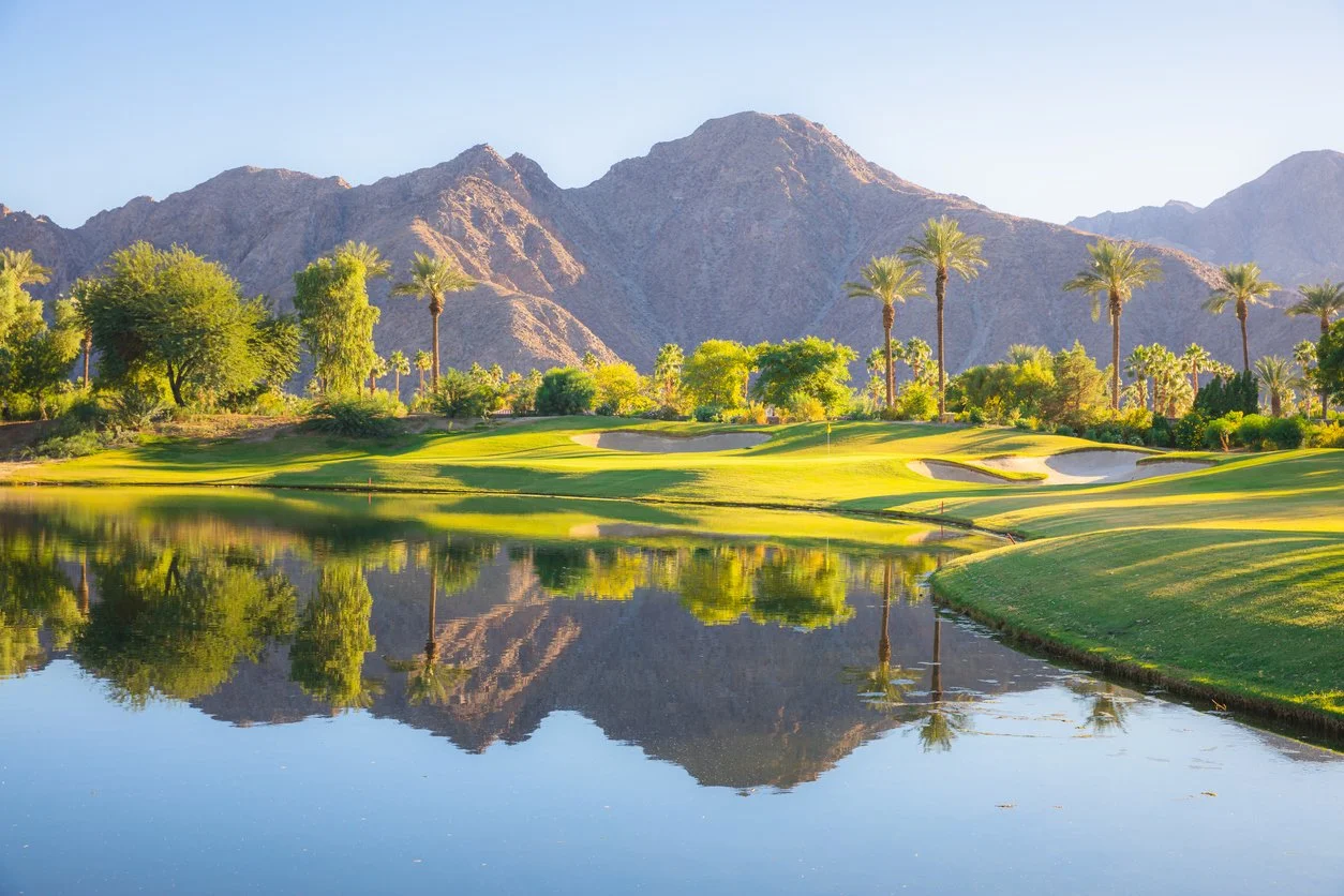 Scenic golf course near mountains, with water reflecting trees and landscape, sunny weather.