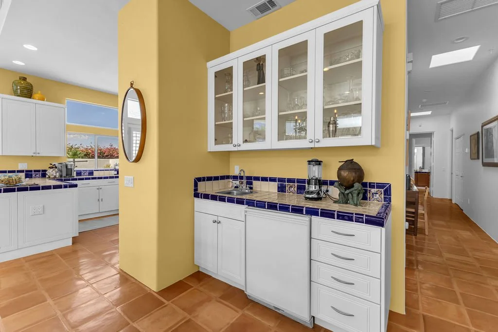 Kitchen with yellow walls, white cabinets, blue tile counter, and terracotta floor, featuring a small sink area and a hallway in the background.