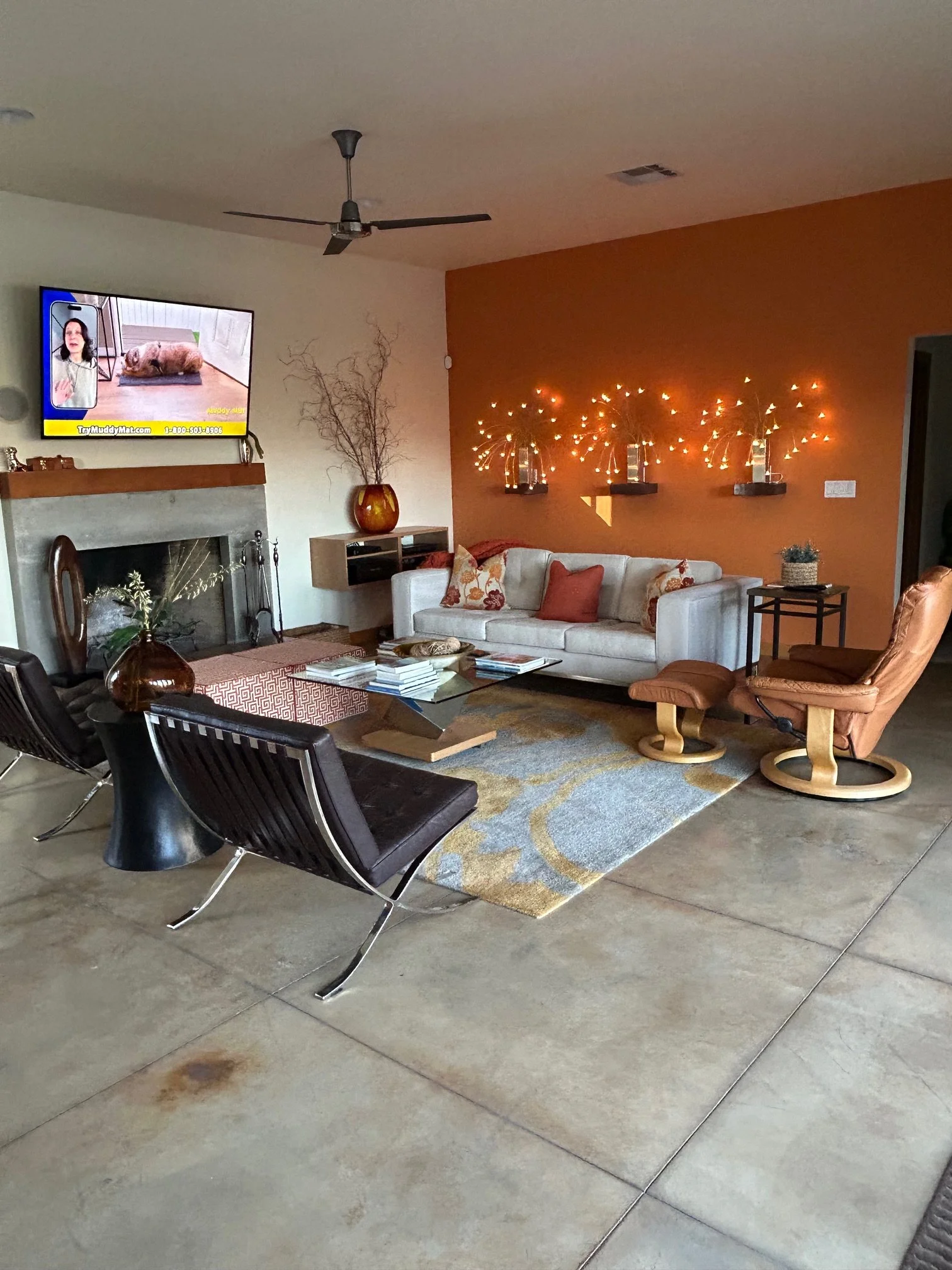 Living room with a fireplace, television, white couch with pillows, decorative wall art, and black chairs around a glass coffee table, with a gray concrete floor.