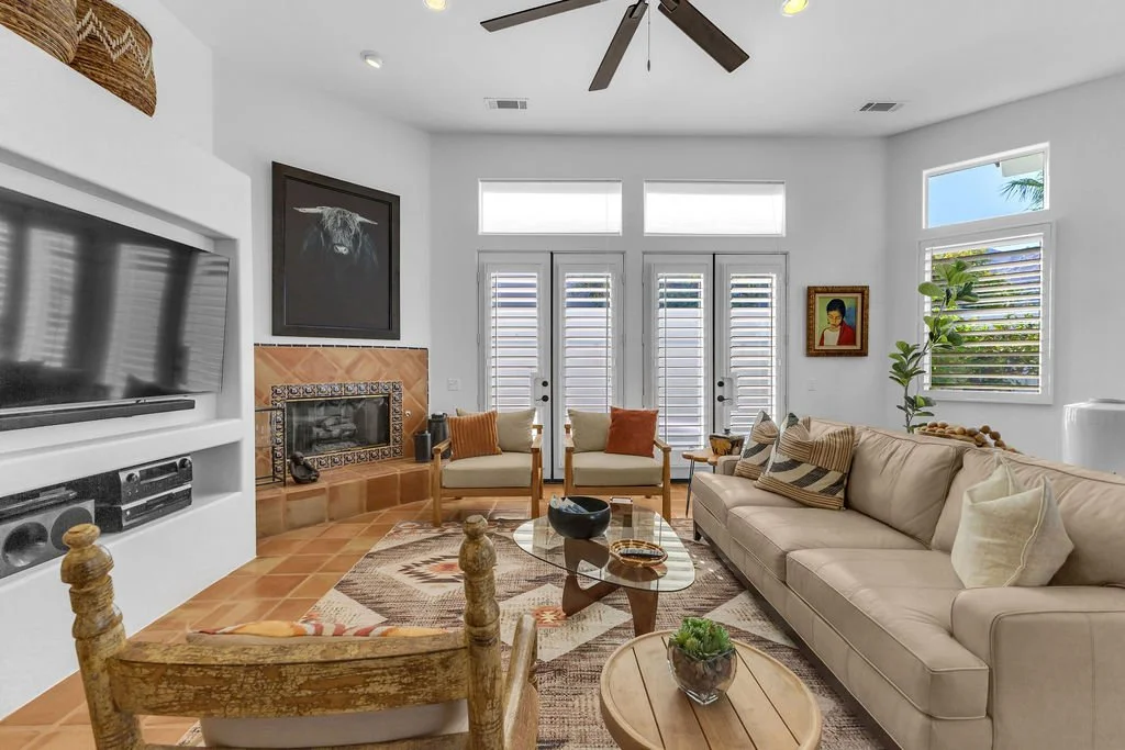 A bright living room with a cream sofa, two cream armchairs, a wooden coffee table, and a colorful area rug. The room has a fireplace with decorative tiles, a large flat-screen TV, and several windows with blinds. There are framed artworks on the wal