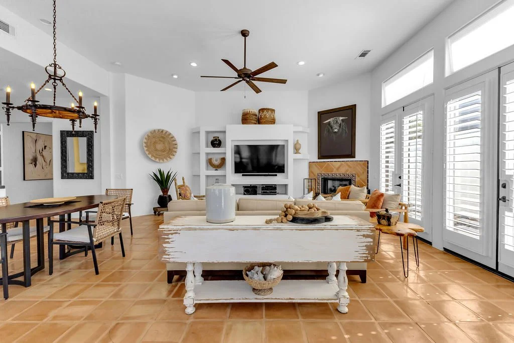 Living room with white walls, farmhouse furniture, a flat-screen TV, a ceiling fan, a large dining table, and front doors with shutters.