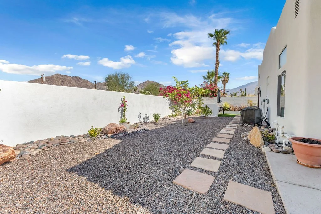A backyard patio with a stone pathway, desert plants, and a white wall, with mountains and a blue sky in the background.
